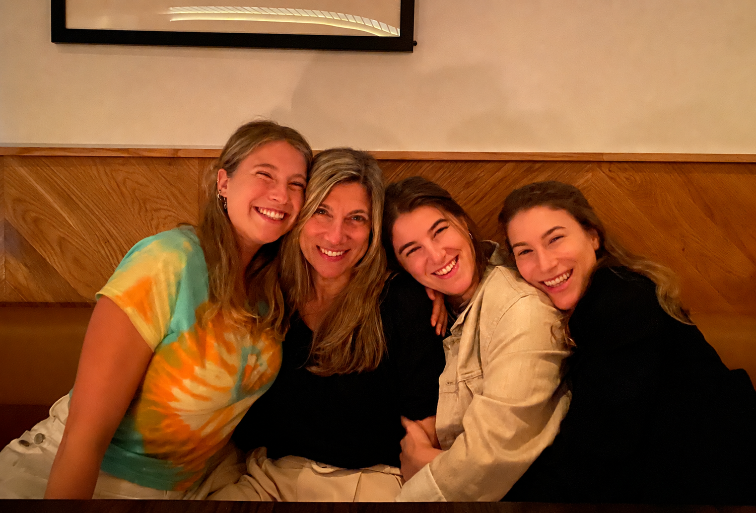 Four women smiling and embracing each other in a warm indoor setting.