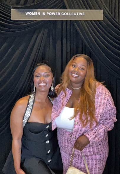 Two women smiling at an event, standing in front of a black curtain with a sign that reads 'Women in Power Collective'.