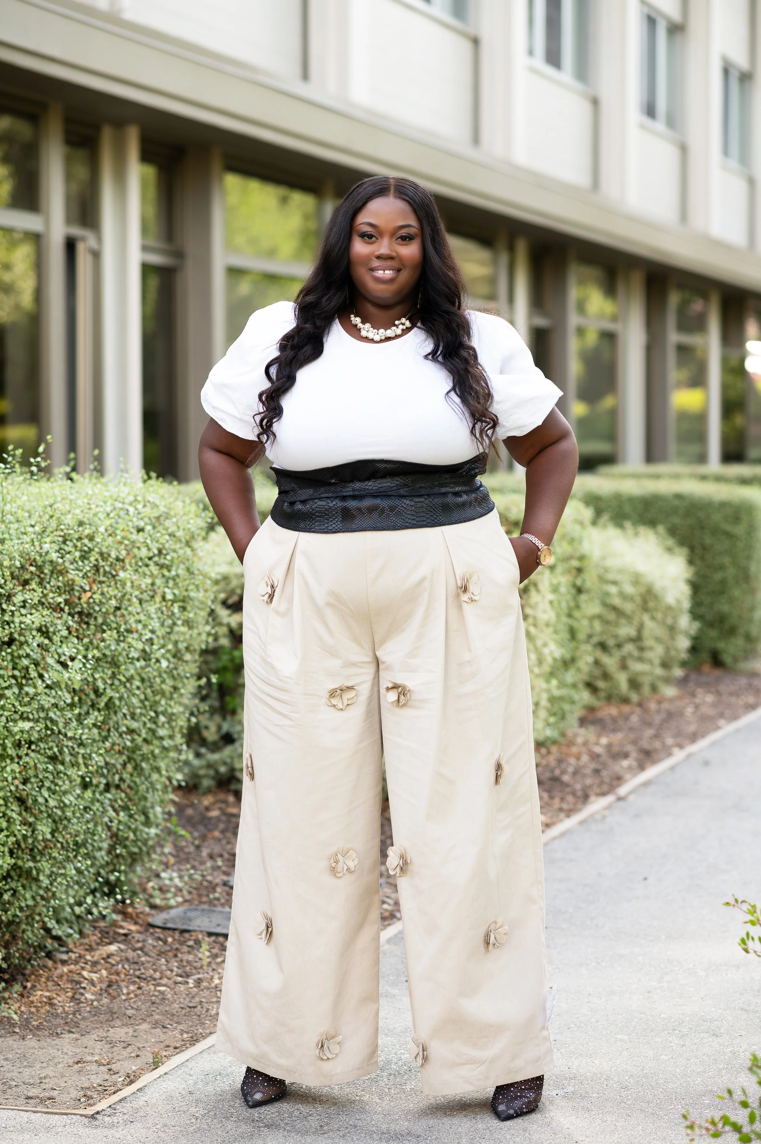 A woman standing outside next to a low hedge, wearing a white top, cream wide-leg pants with floral embellishments, and black high heels, with her hands in her pockets and smiling.