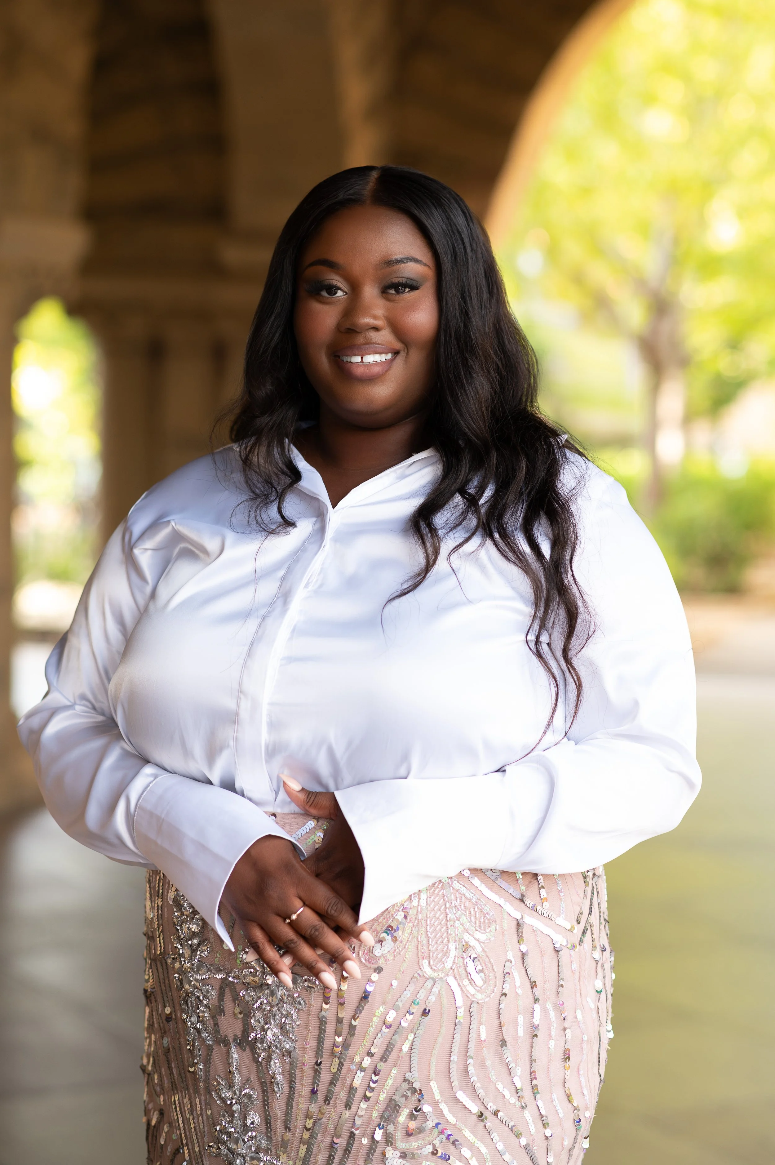 A woman with long black wavy hair wearing a white blouse and a beaded, patterned skirt standing under a stone bridge in a park.