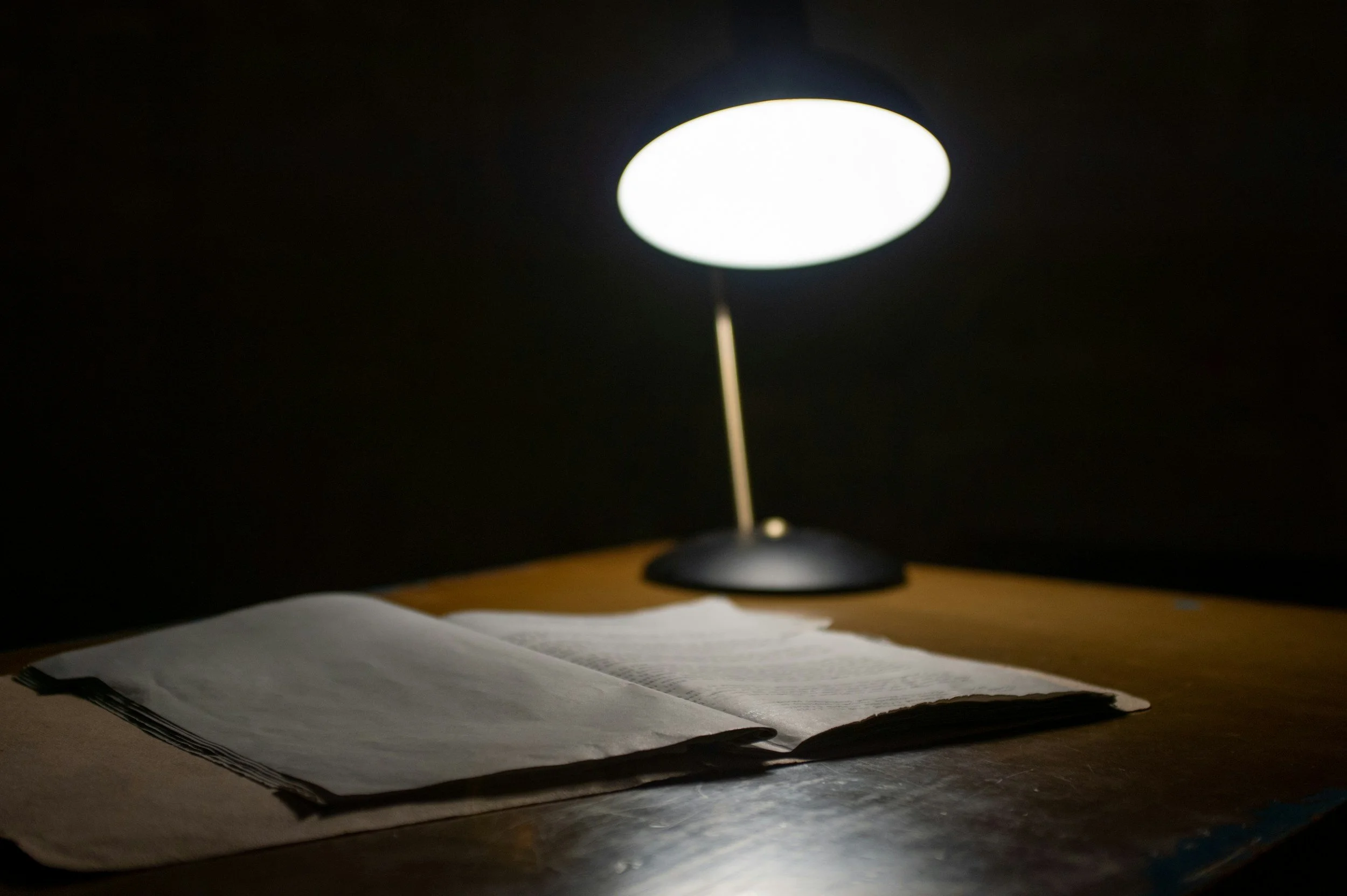 A desk lamp illuminating an open book on a wooden surface in a dark room.