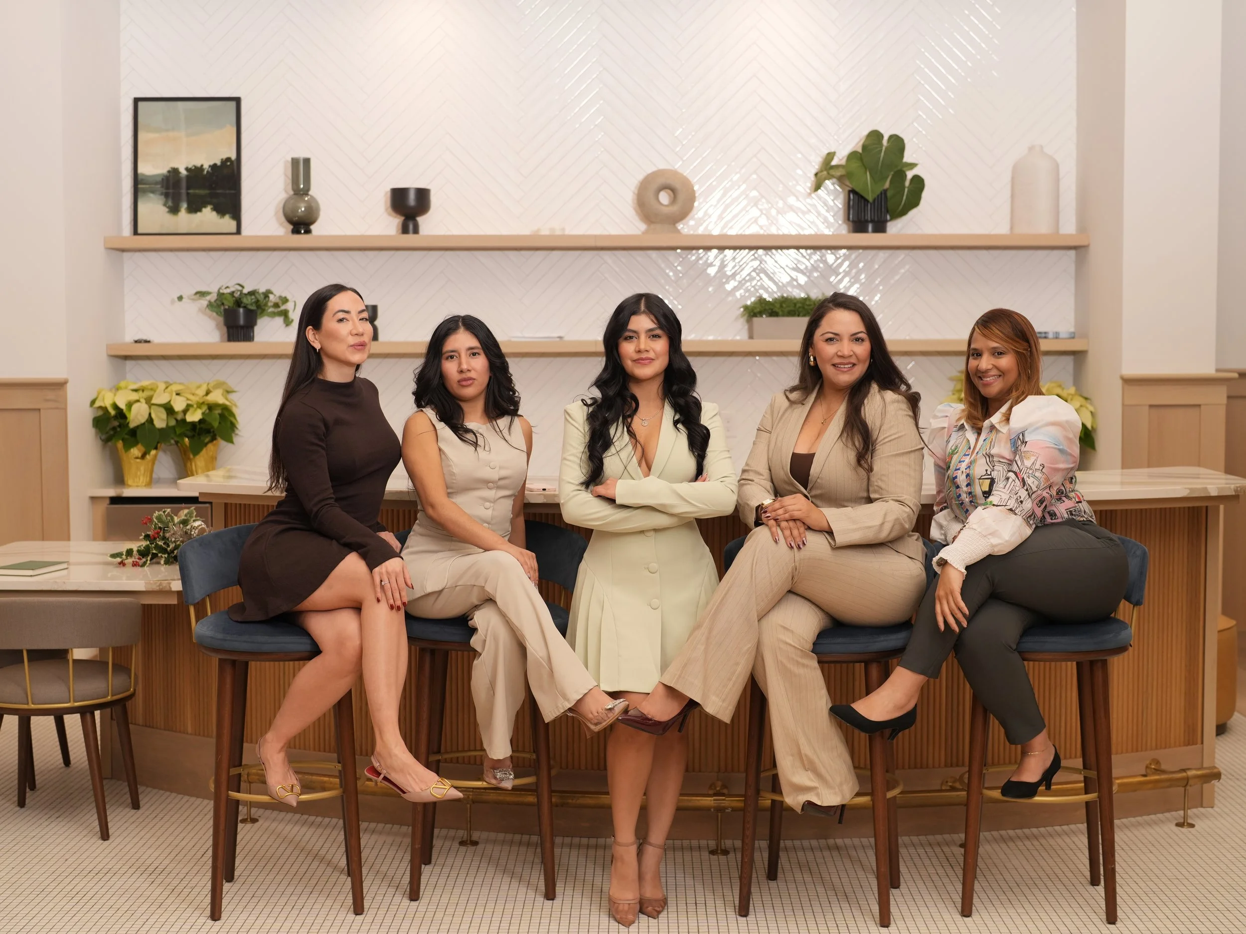 Cinco mujeres sentadas en una barra de un espacio decorado con plantas y cerámicas, en tonos neutros y azul.