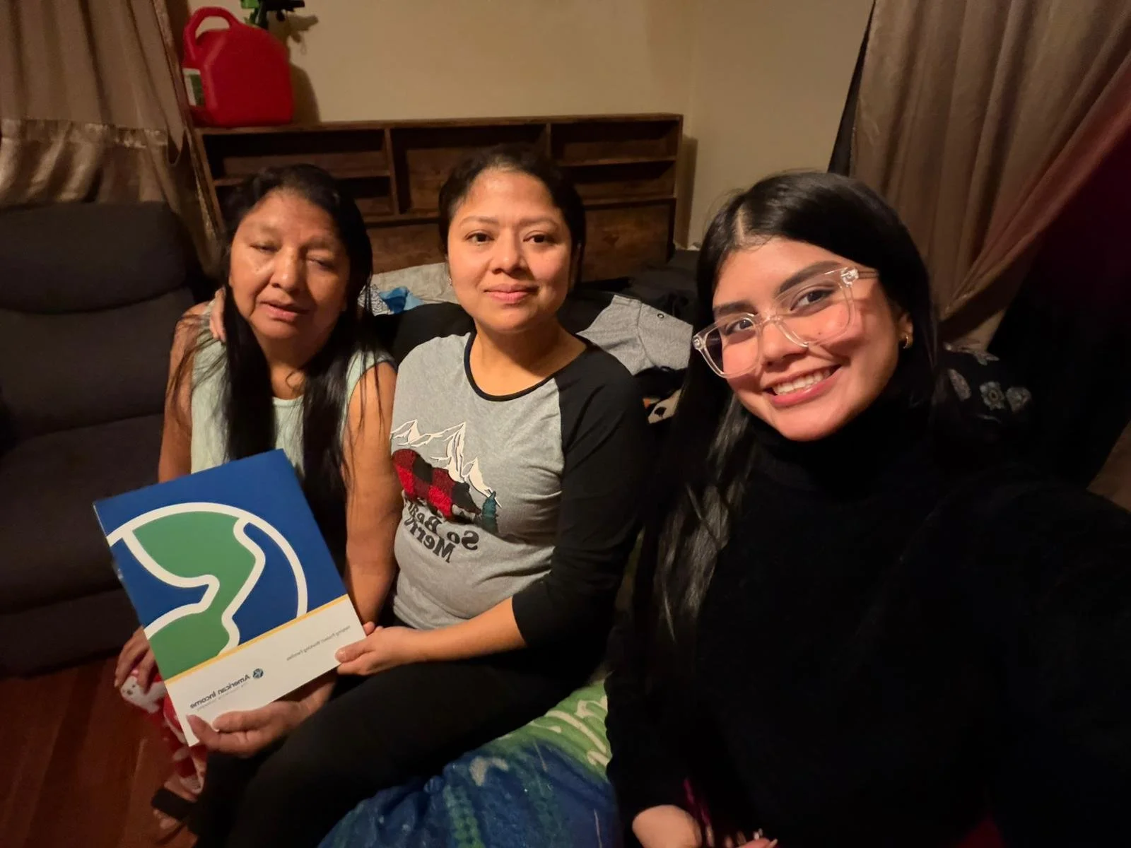 Tres mujeres en un sofá, una de ellas sosteniendo un folleto con un logo y texto, en una sala con cortinas y muebles de madera.