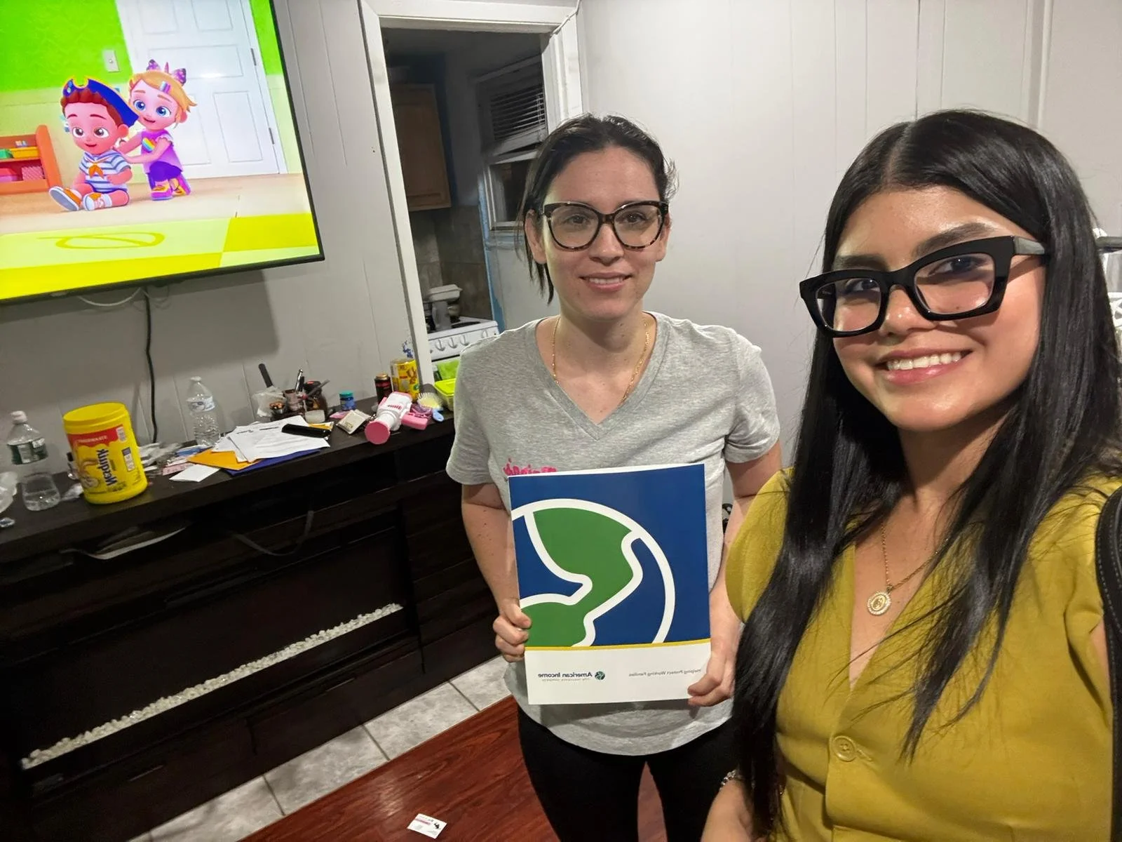 Dos mujeres sonrientes posando para una selfie en una habitación, una de ellas sostiene un folleto con un logotipo de un camino y un árbol, detrás hay un televisor que muestra animaciones infantiles, y una mesa con varios objetos como botellas, medic