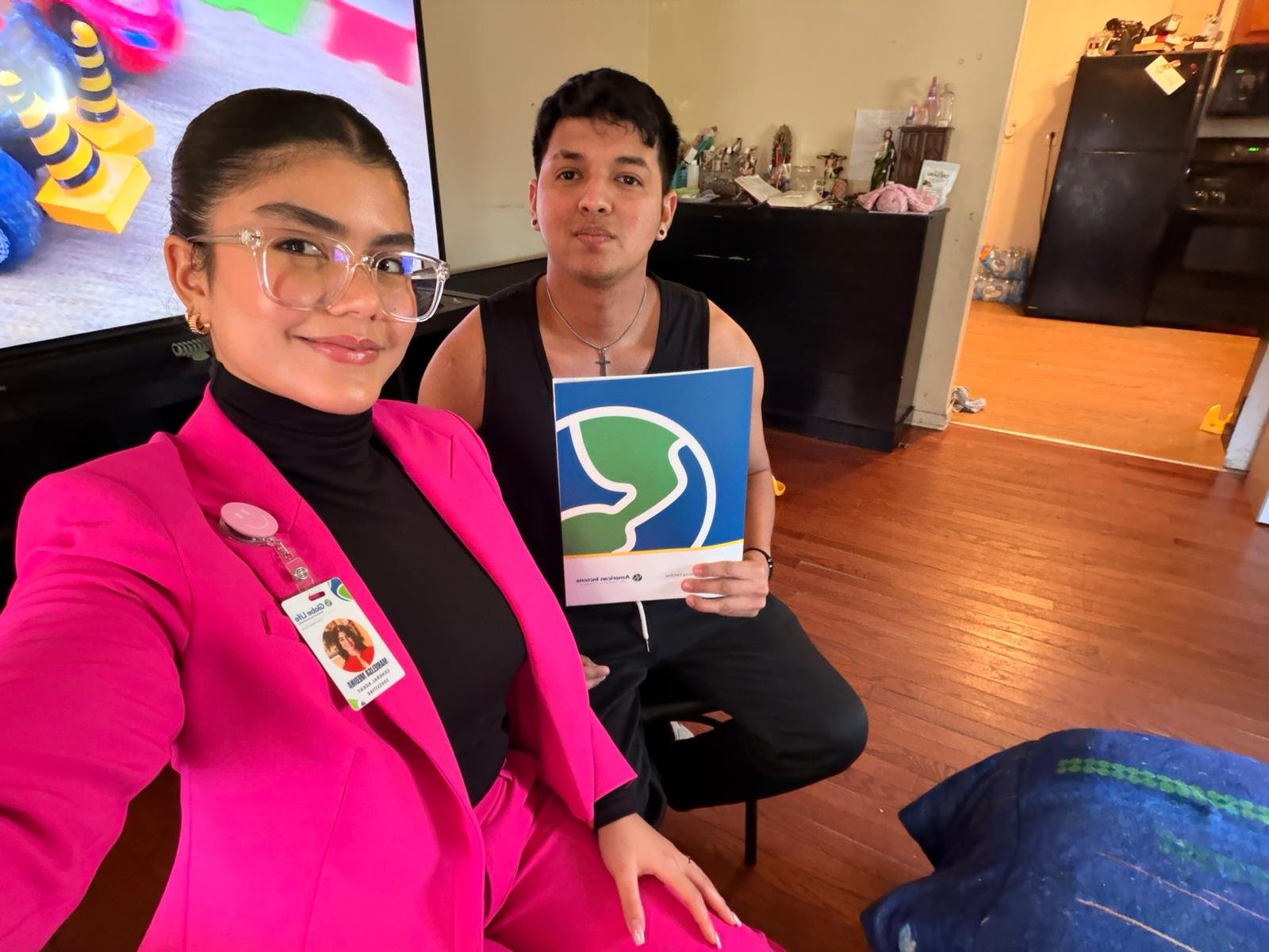 Dos personas, una mujer con traje rosado y gafas, y un hombre con camiseta sin mangas, posando en una sala, ella lleva una identificación y él sostiene un documento con un logo con un dibujo de un árbol y un fondo azul y verde.