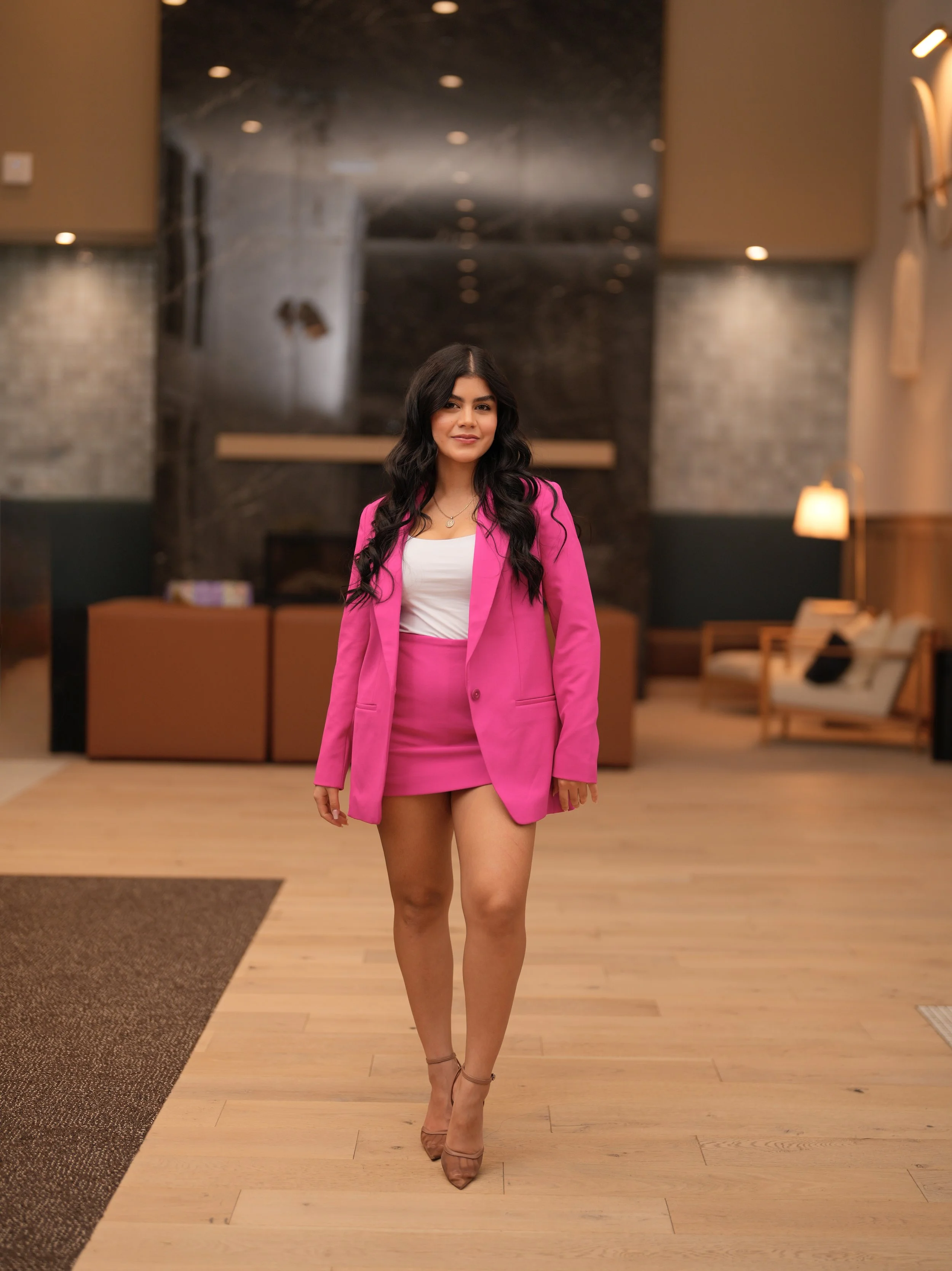 Mujer con cabello largo y oscuro, vistiendo un conjunto de saco y falda rosa, caminando en un vestíbulo interior con paredes de ladrillo, luces cálidas y muebles.