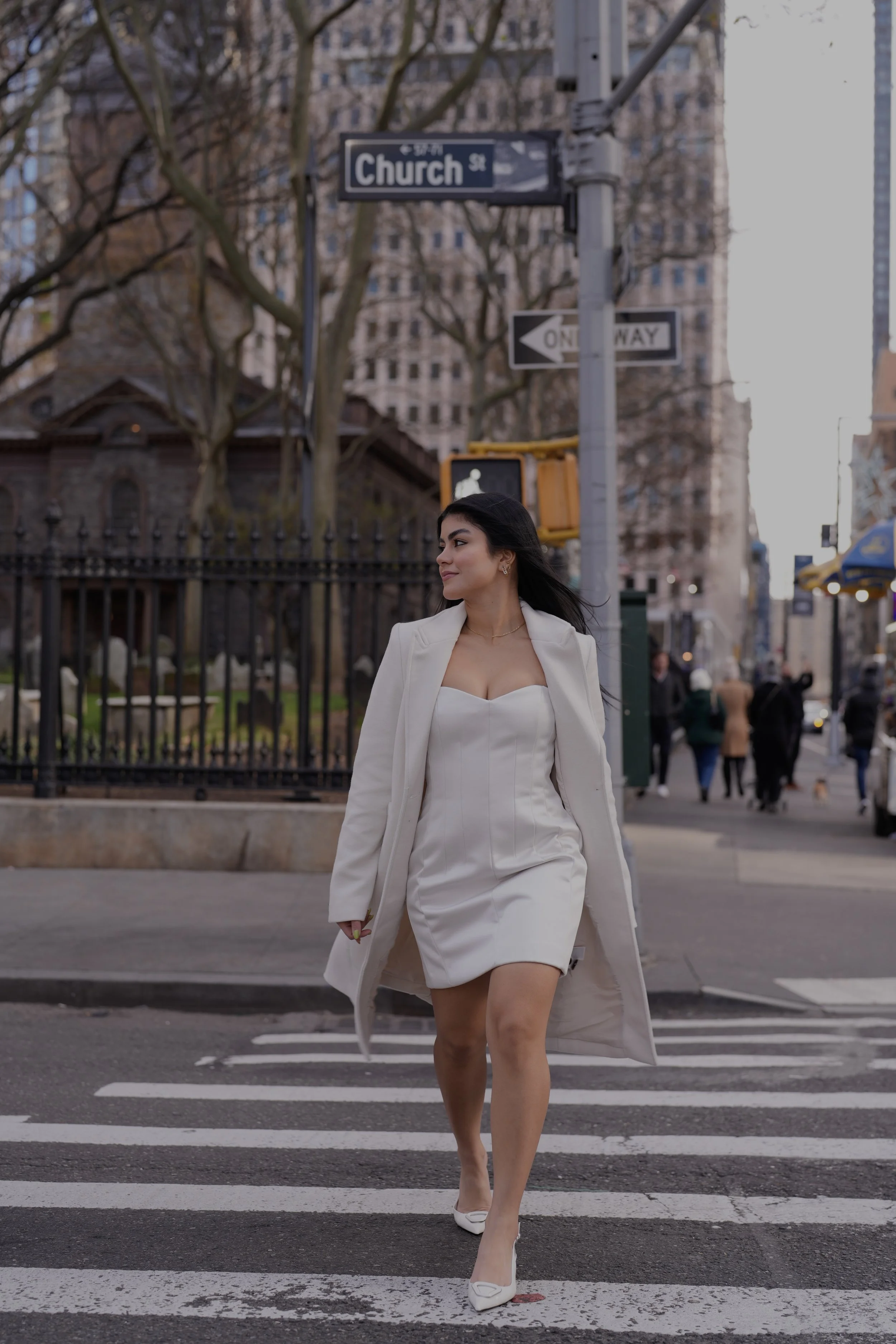 Mujer vestida con vestido y abrigo blancos cruzando la calle en la ciudad.