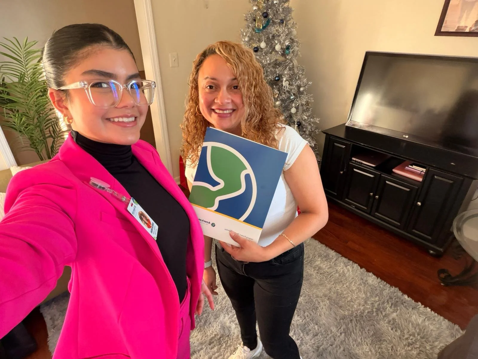 Dos mujeres sonriendo y posando para una selfie en una sala con árbol de Navidad decorado. Una de ellas lleva gafas y un blazer rosa, la otra sostiene un folleto con un logo de un perfil de cabeza en línea verde y azul.