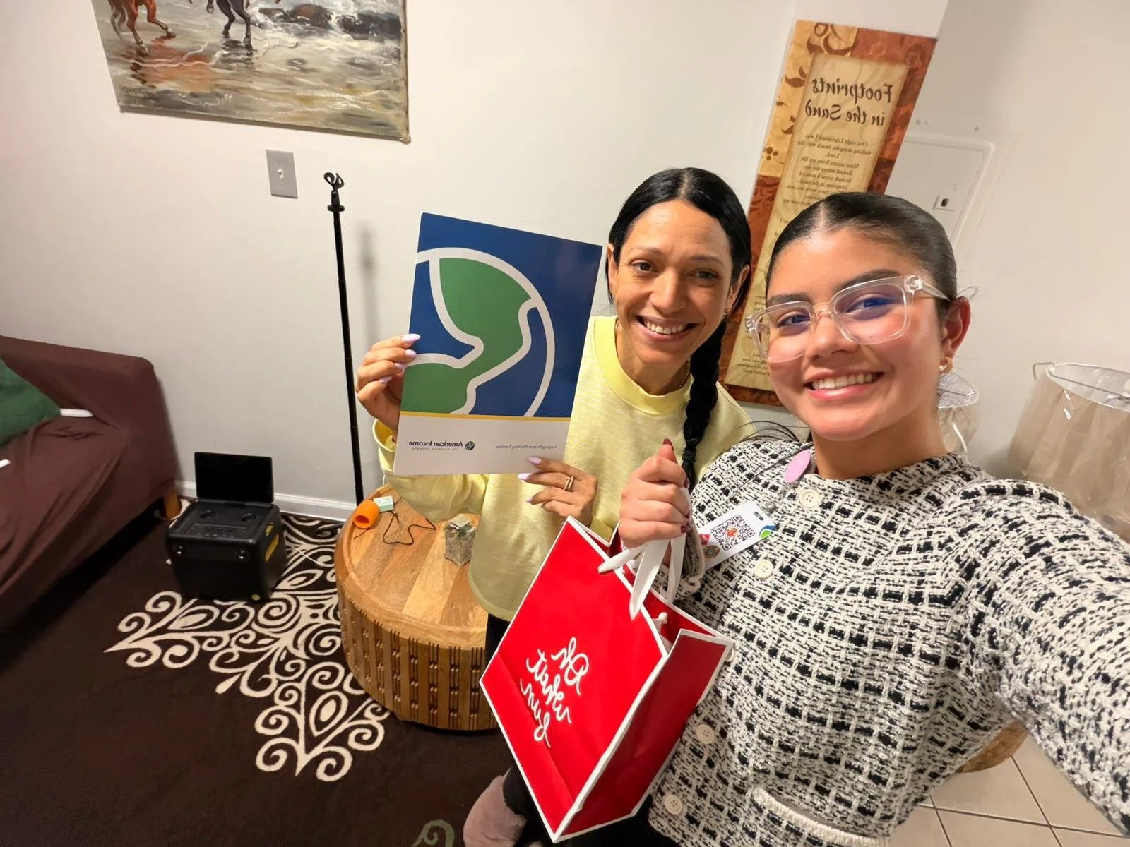 Dos mujeres sonriendo y tomando una selfie en una habitación, una sostiene una bolsa roja y la otra una cartulina con un logotipo azul y verde.