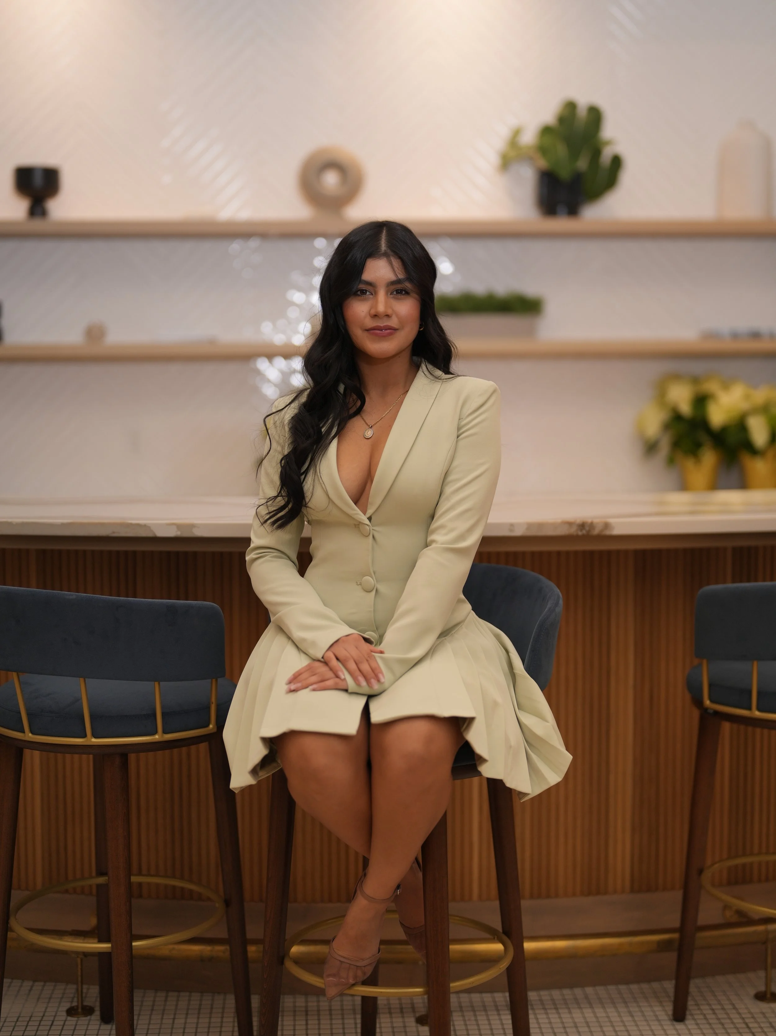 Una mujer con cabello largo y rizado, vestida con un abrigo beige y falda a juego, sentada en una silla en un bar o cafetería con decoraciones modernas y plantas en el fondo.