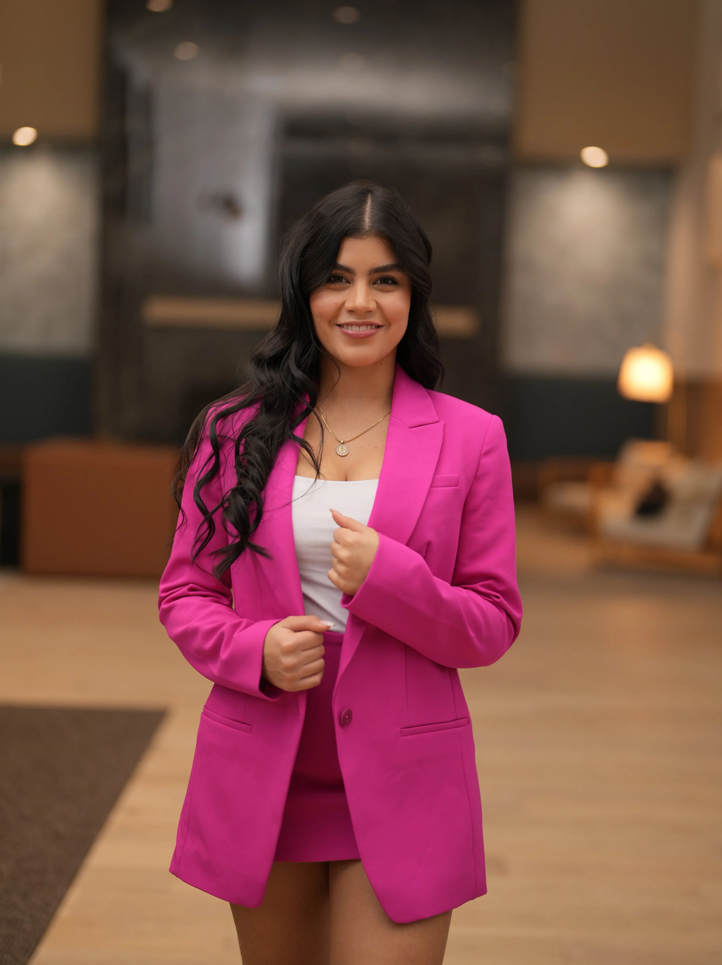 Una mujer sonriendo, con cabello rizado y oscuro, viste un blazer rosa y una blusa blanca, en un ambiente interior con luces cálidas y fondo difuso.