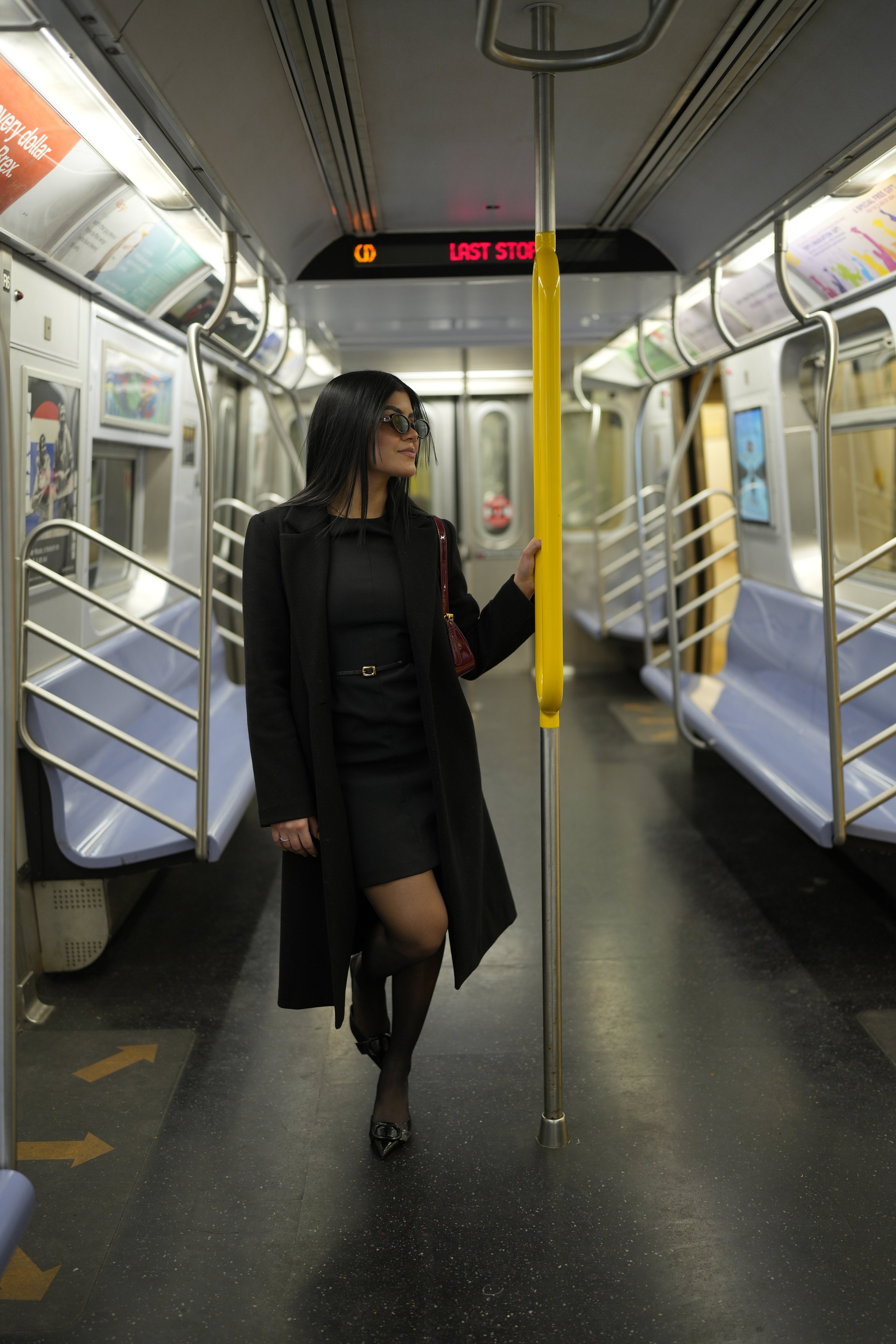 Una mujer vestida de negro en un vagón de metro, sosteniendo una barra amarilla con una mano, con un fondo de asientos azules y anuncios en las paredes.
