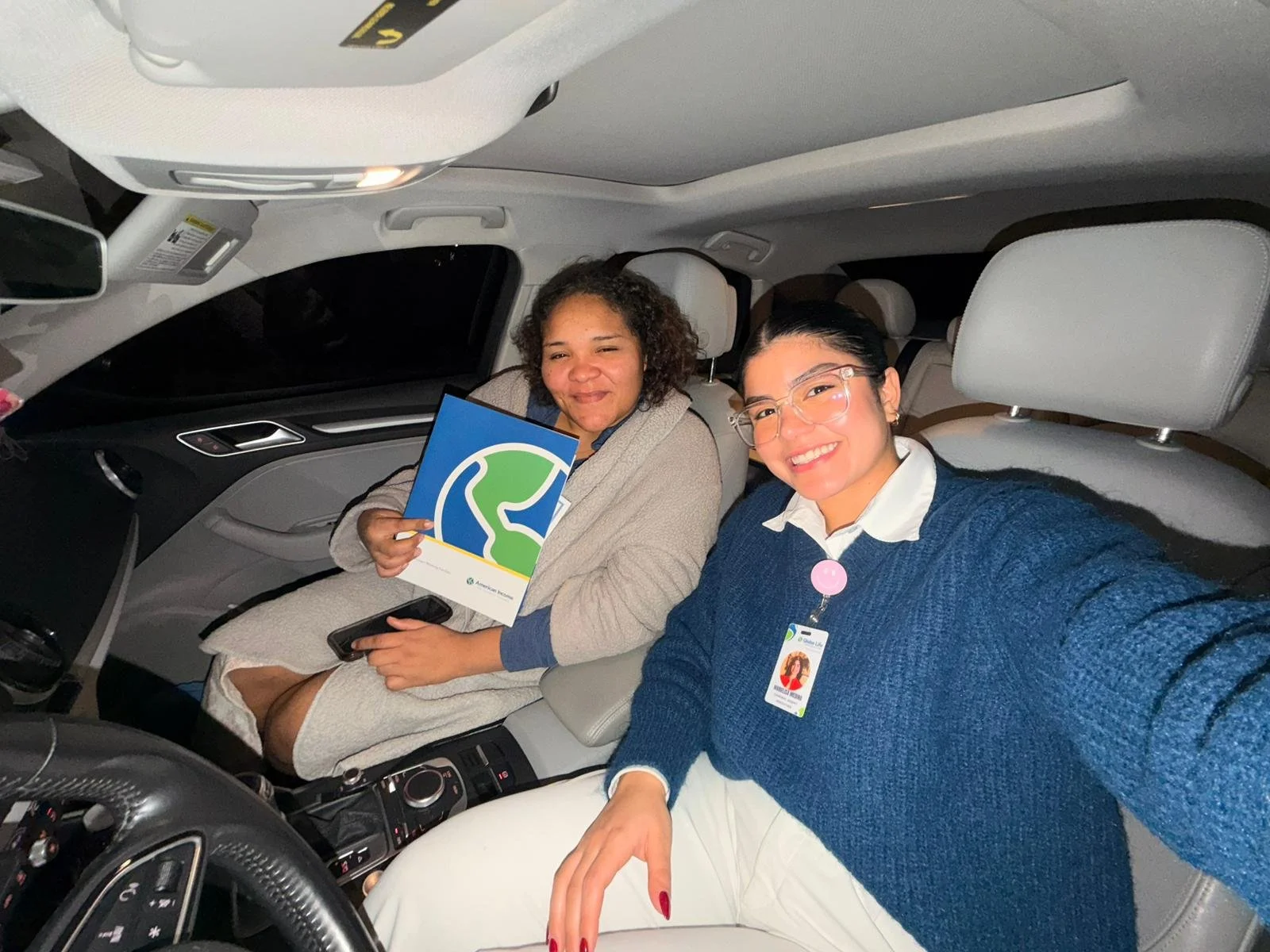 Dos mujeres sonrientes sentadas en un coche, una con gafas, usando camisa blanca y suéter azul, la otra con cabello rizado y piel morena, sosteniendo un folleto con un logo de un árbol verde y azul.