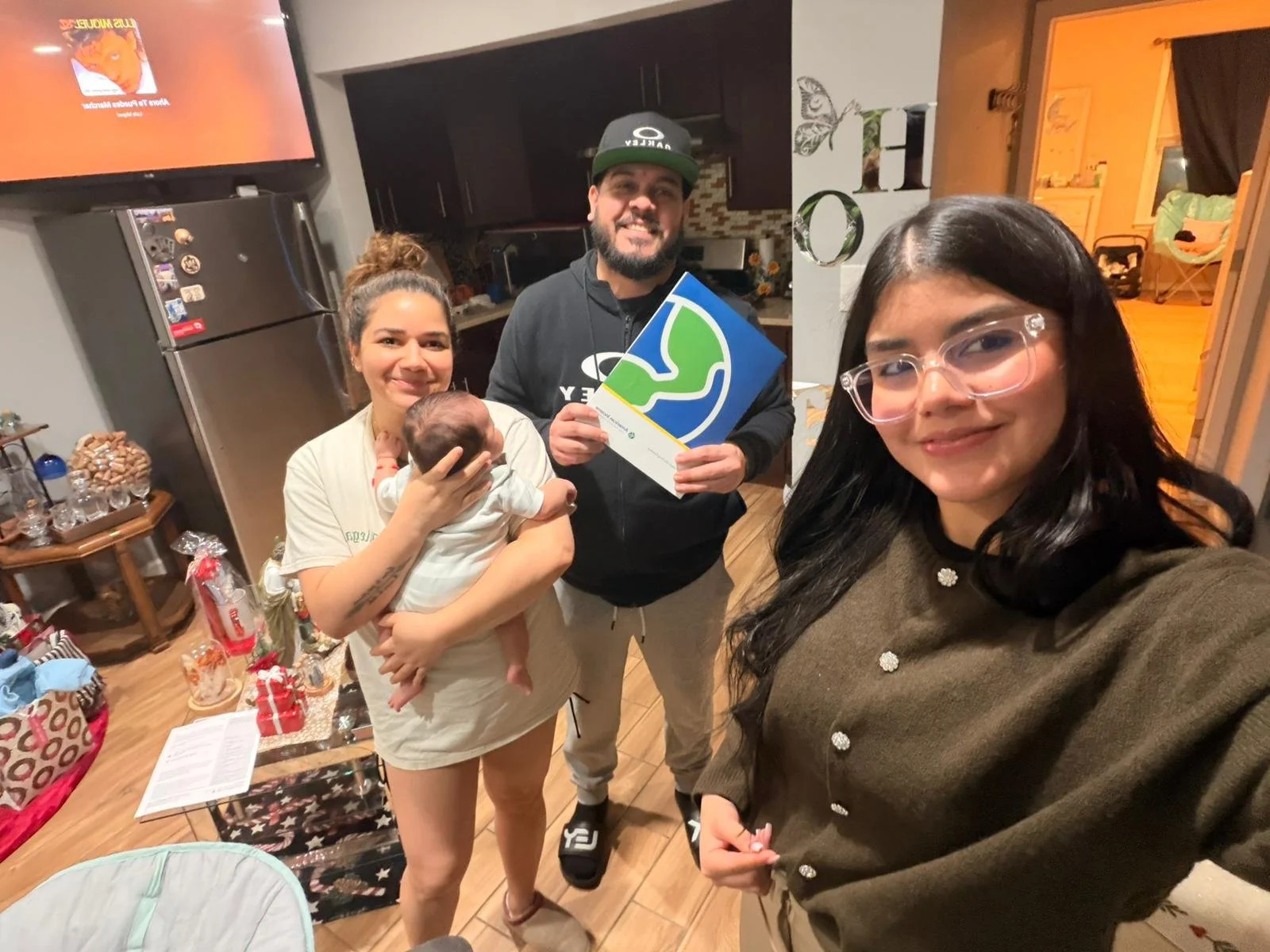 Cuatro personas en una reunión en casa, una mujer sostiene un bebé, otra mujer está tomando la selfie, un hombre tiene un diploma, en la sala y cocina de fondo.
