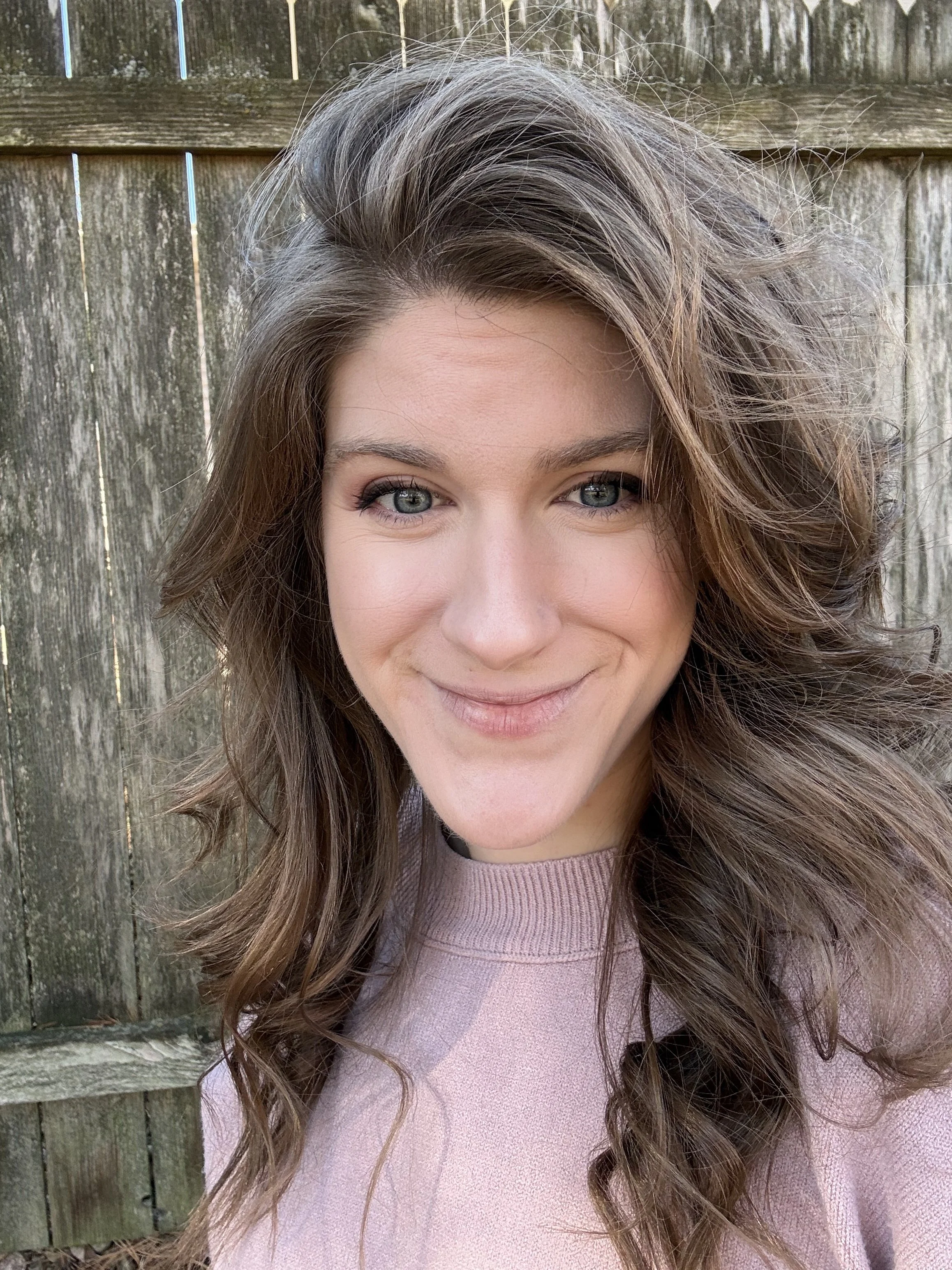 Close-up of a woman with wavy brown hair, blue eyes, and a soft smile, standing outdoors in front of a wooden fence.