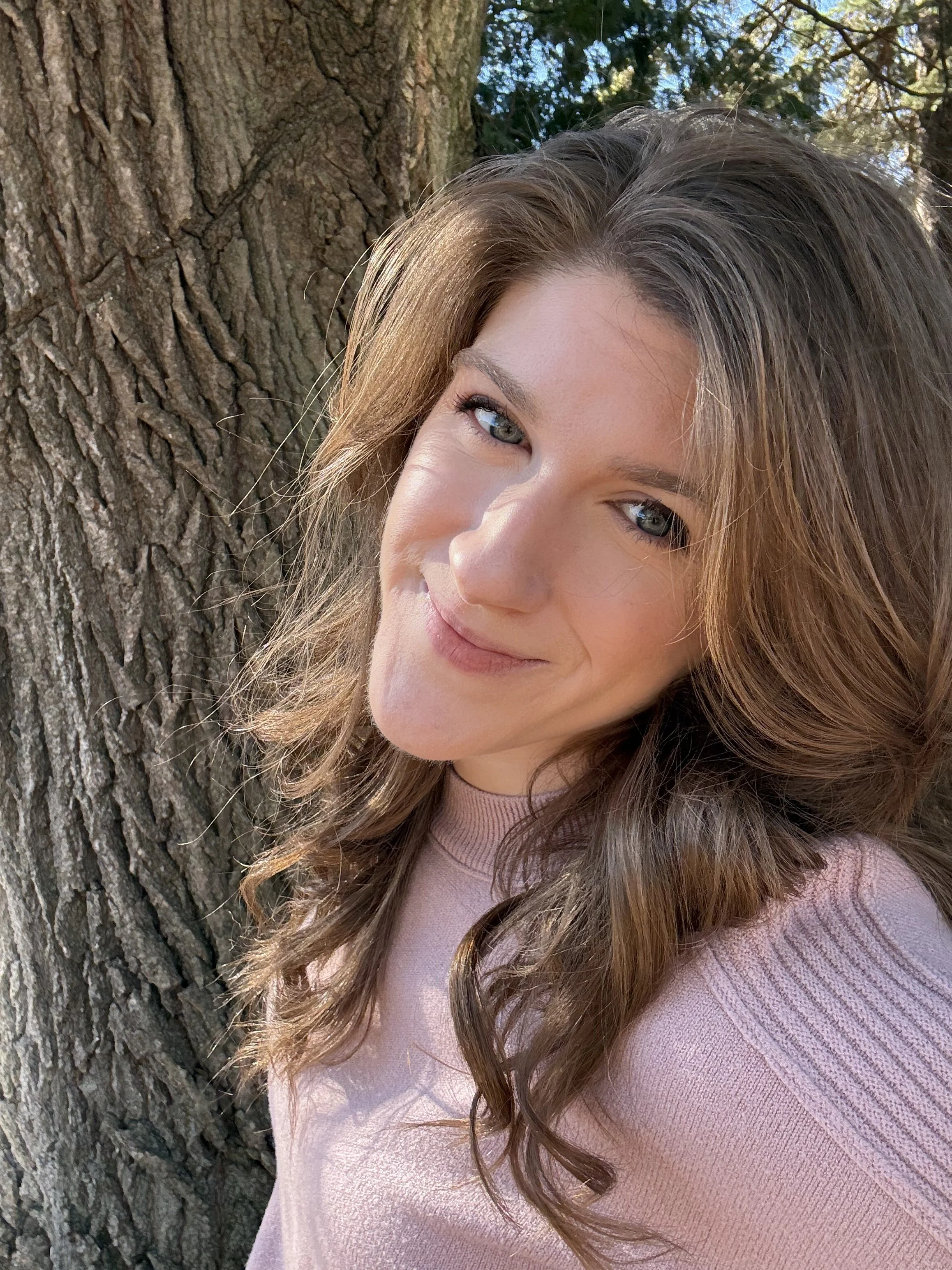 A young woman with light brown, wavy hair and blue eyes smiling while leaning against a large tree outdoors on a sunny day.