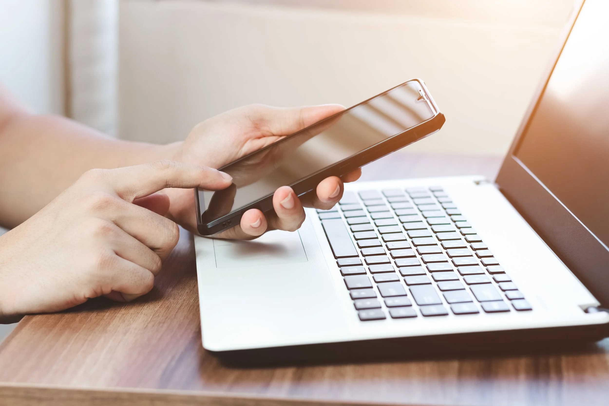 Person holding a smartphone above a laptop on a wooden desk.