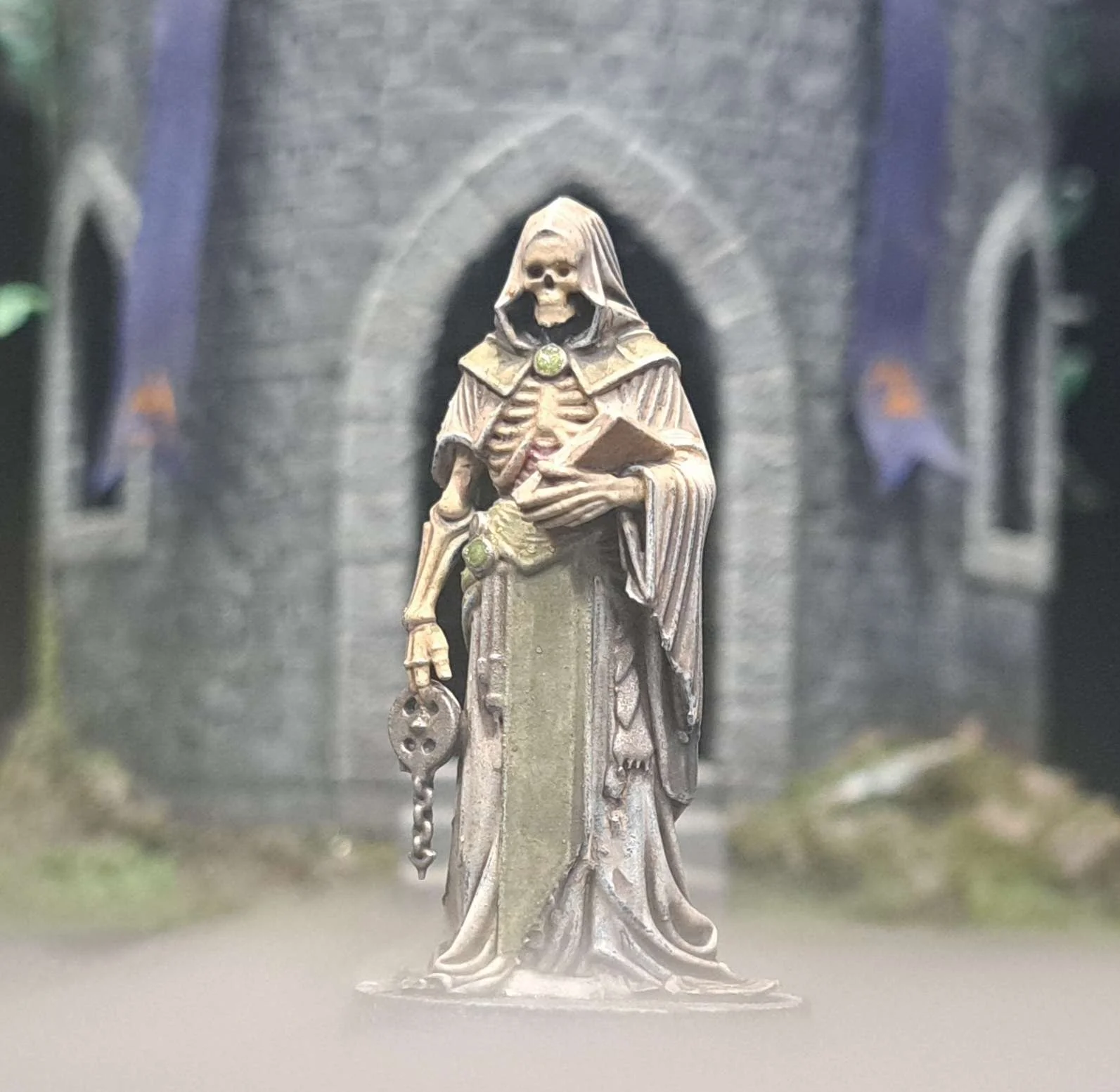Miniature figurine of a skeleton dressed as a dark wizard, holding a book in one hand and a staff with a key in the other, standing in front of a castle with banners.