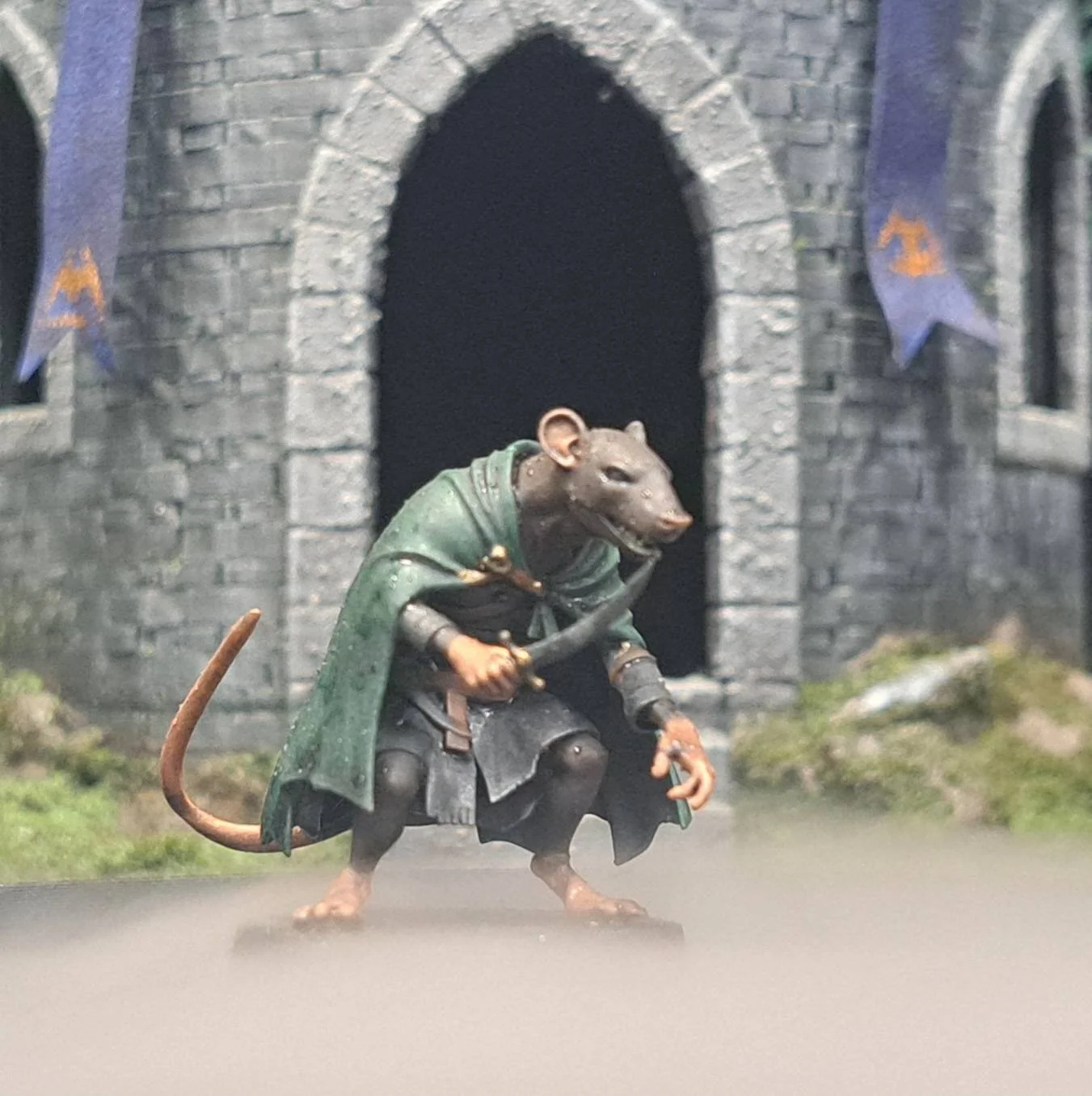A detailed figurine of a mouse dressed as a medieval knight, wearing a green cloak and holding a sword, standing in front of a stone castle archway with blue flags.