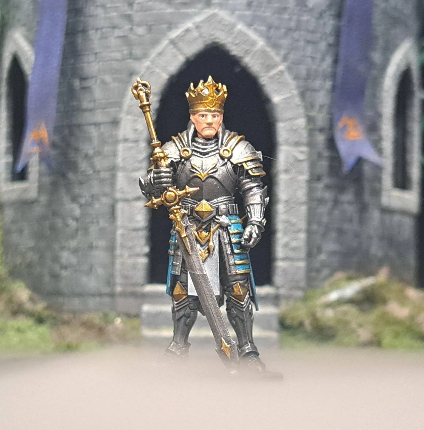 A figure dressed as a medieval king in black and gold armor, wearing a crown, holding a sword, standing in front of a stone castle entrance.
