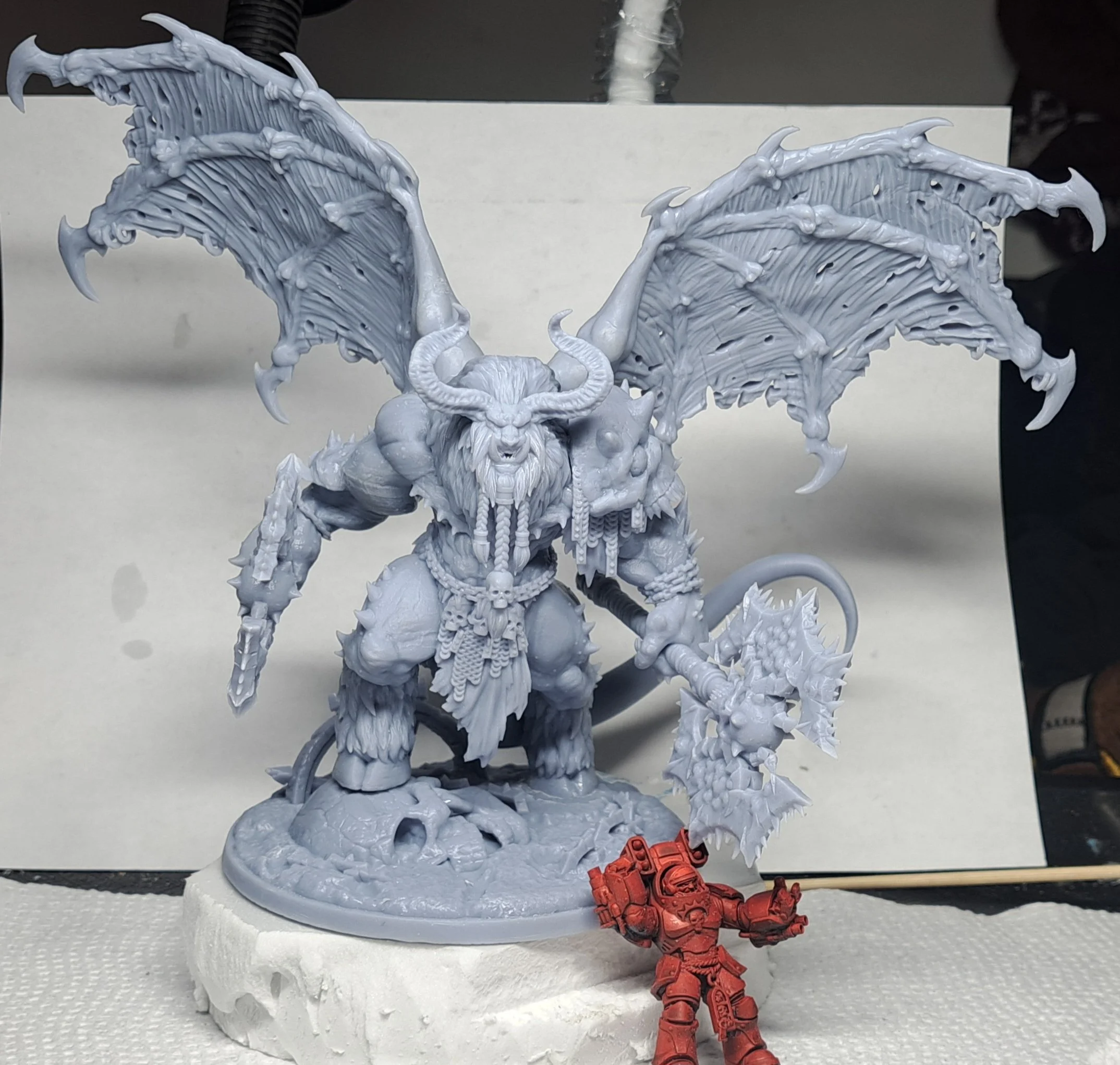 A detailed miniature figurine of a winged demon or monster with horns, a beard, and muscular build, standing on a textured base. It wields a large, spiked weapon and is adorned with skulls and chains. A small red miniature of a space marine in armor is in front of it.