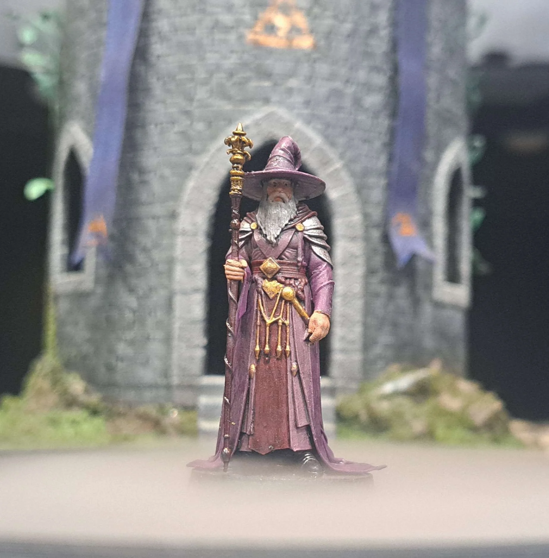Miniature wizard figurine in purple robes and hat, holding a staff, standing in front of a medieval style miniature castle with arched windows and a stone facade.