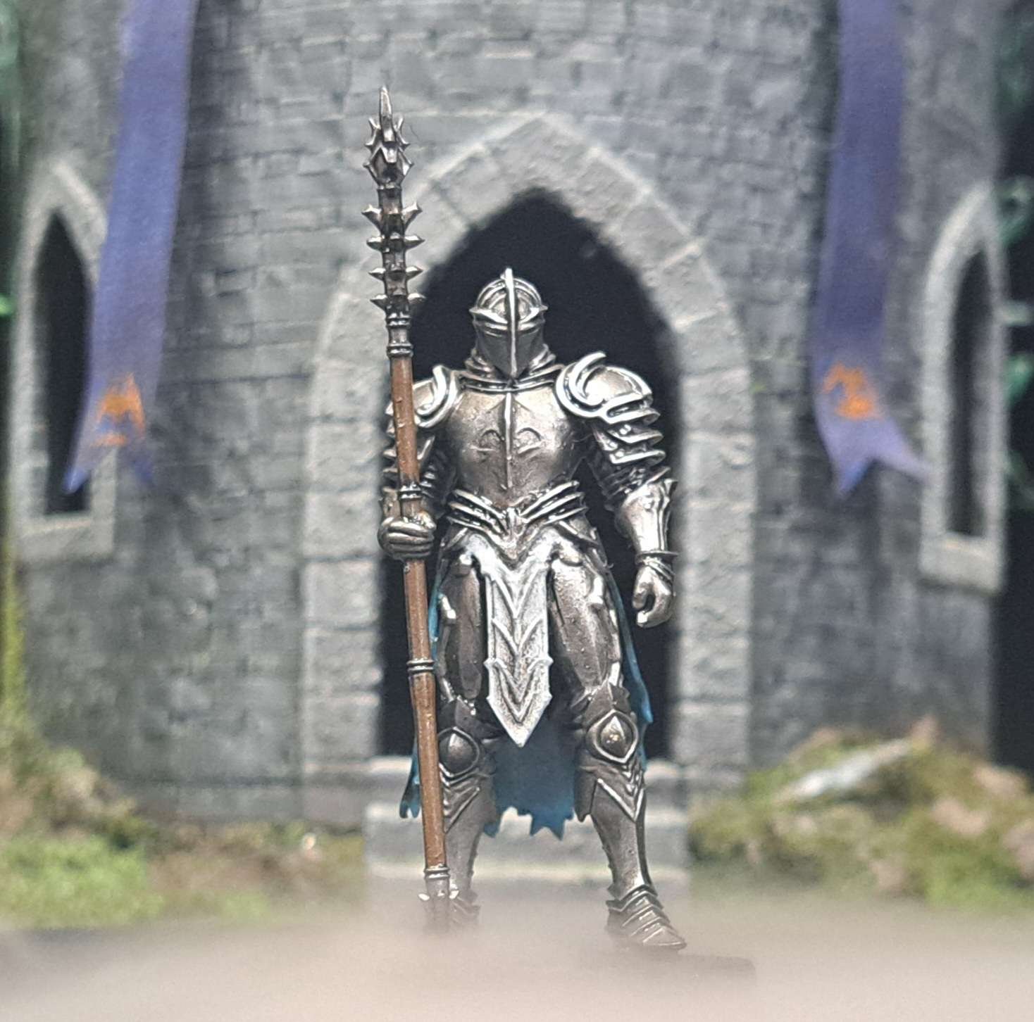 Miniature figurine of a knight in metallic armor holding a long spear, standing in front of a castle entrance with stone walls and arched doorway, decorated with banners.