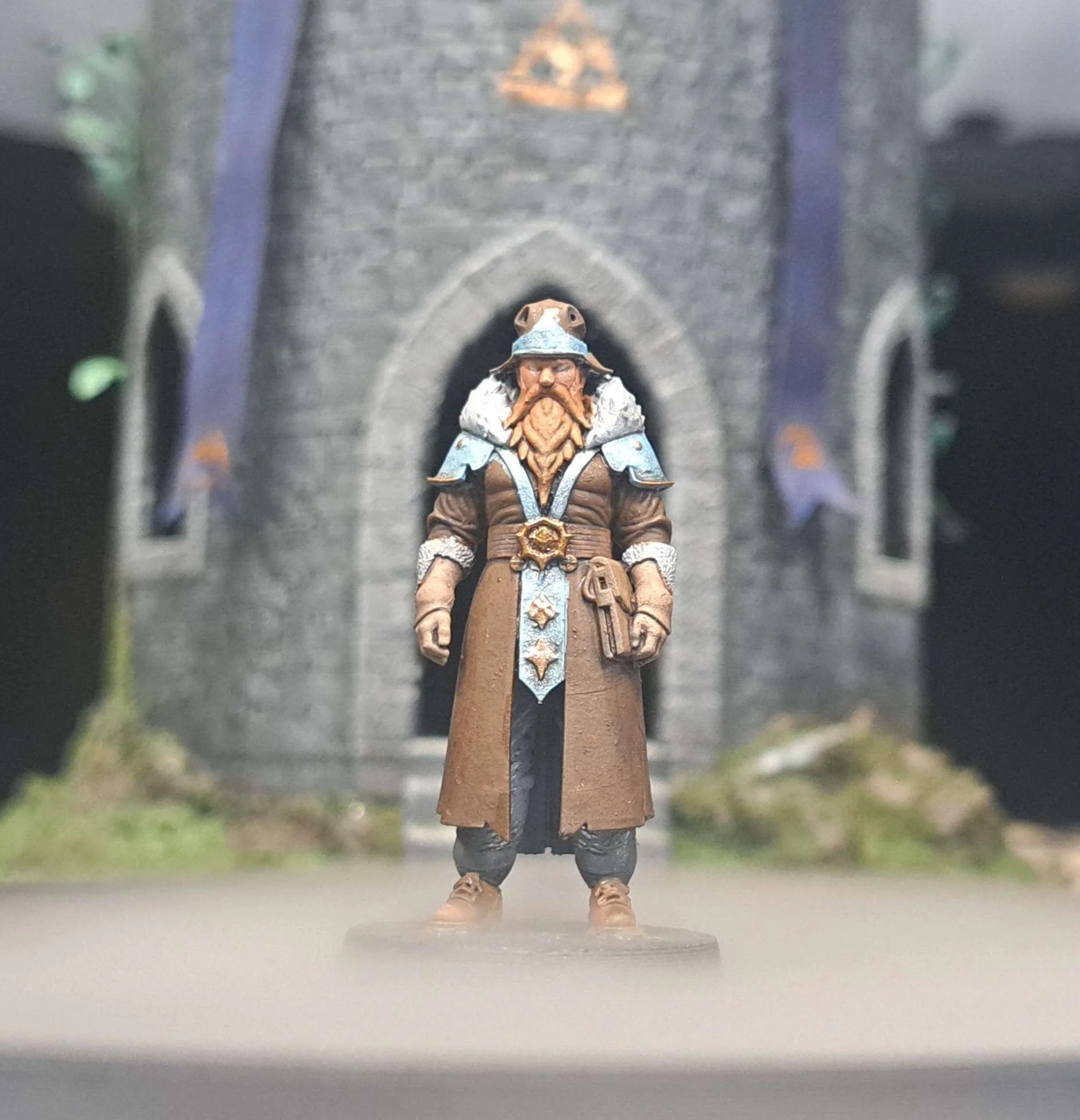 A detailed miniature figurine of a dwarf warrior with a long beard, wearing armor and a long coat, standing in front of a model castle wall.