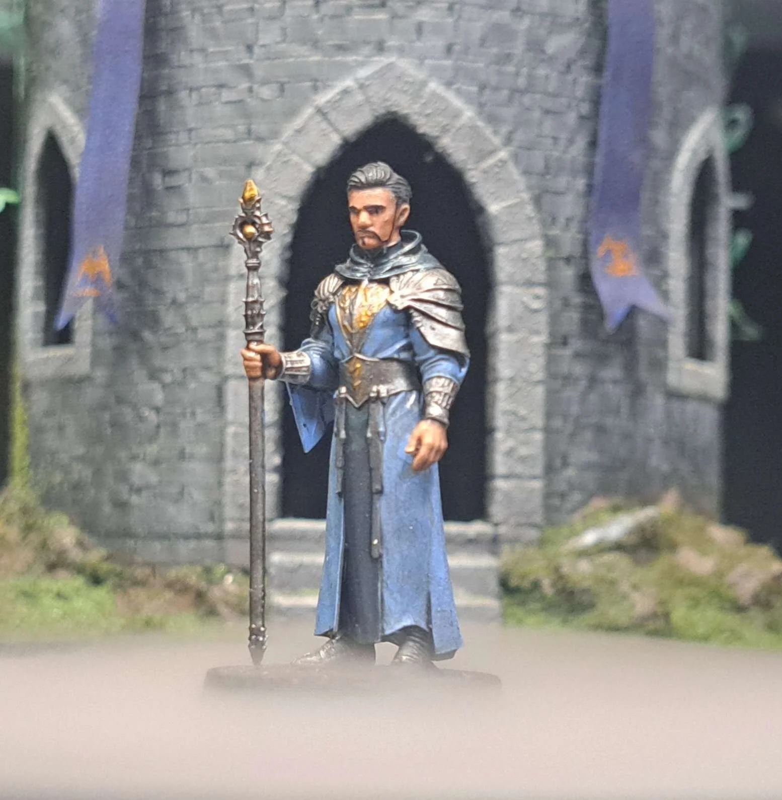 Miniature figurine of a medieval knight in blue robes holding a staff, standing in front of a stone castle entrance with banners.