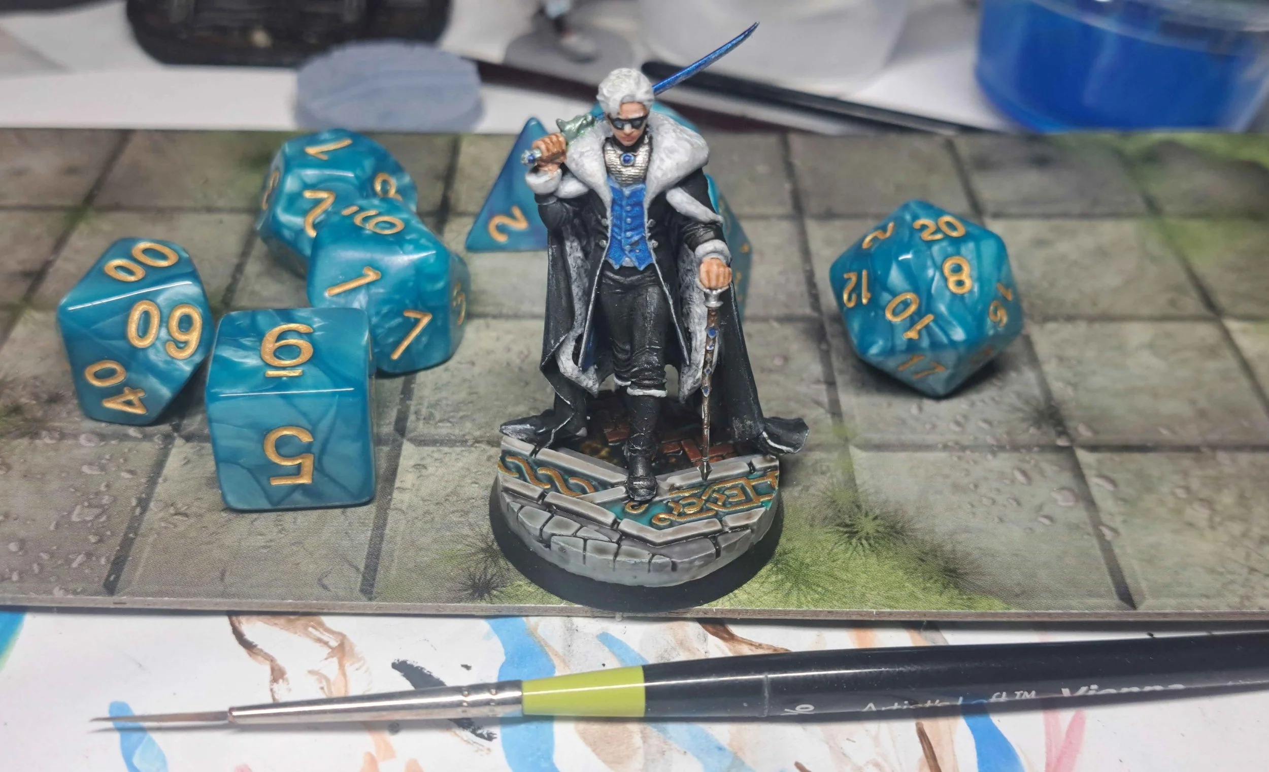 Tabletop gaming scene with a fantasy miniature figure of an elderly wizard with white hair, dressed in a blue and black robe, holding a staff and sword. Surrounding the figure are three blue polyhedral dice showing various numbers, on a textured game mat with a painted grid and grassy details. A paintbrush is lying in front of the scene.
