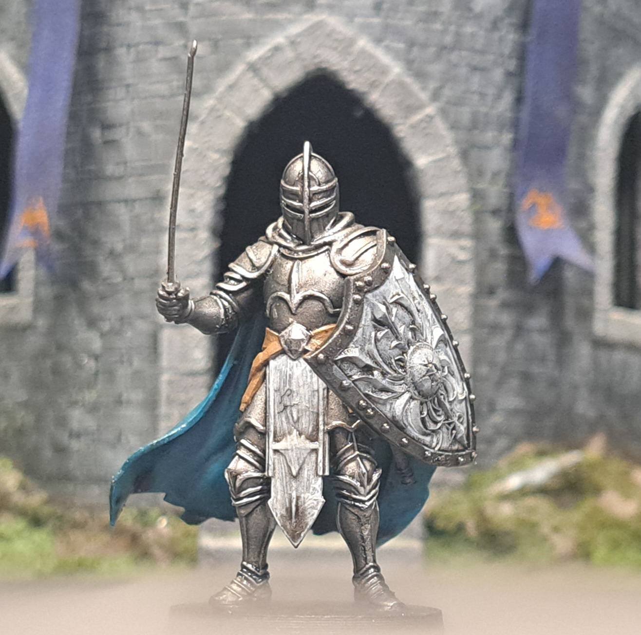 Miniature medieval knight figurine in silver armor holding a sword, with a decorated shield, standing in front of a stone castle backdrop.