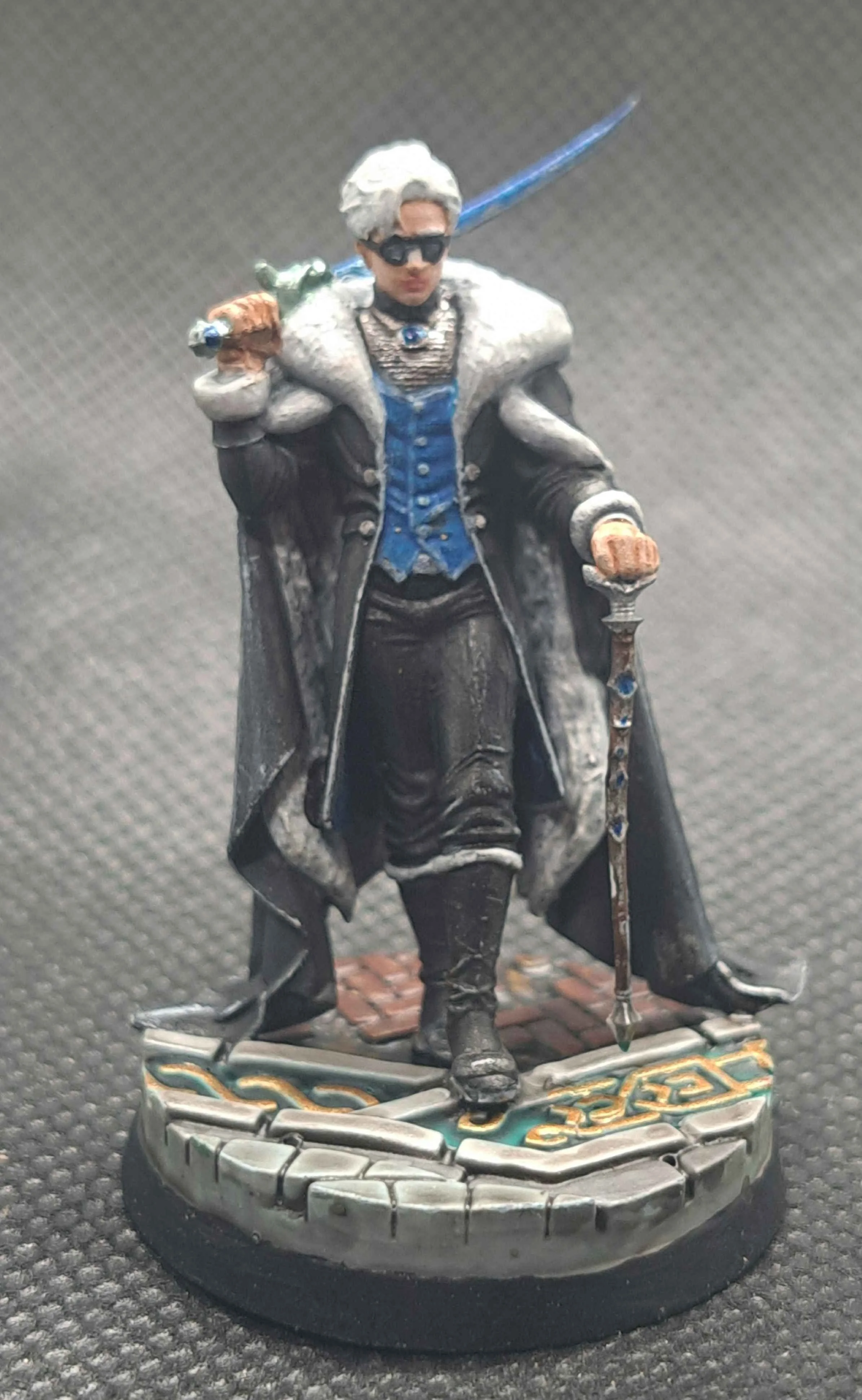 Miniature figurine of a man wearing black sunglasses, a long dark coat with gray fur collar, walking on a round base with stone and metallic details, holding a staff.