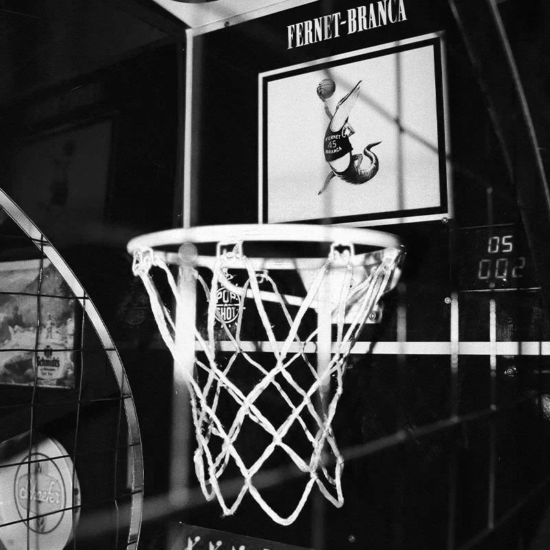 Black and white photo of a basketball hoop with a backboard, labeled 'Fernet-Branca,' and a player silhouette illustration, taken inside a bar or arcade.