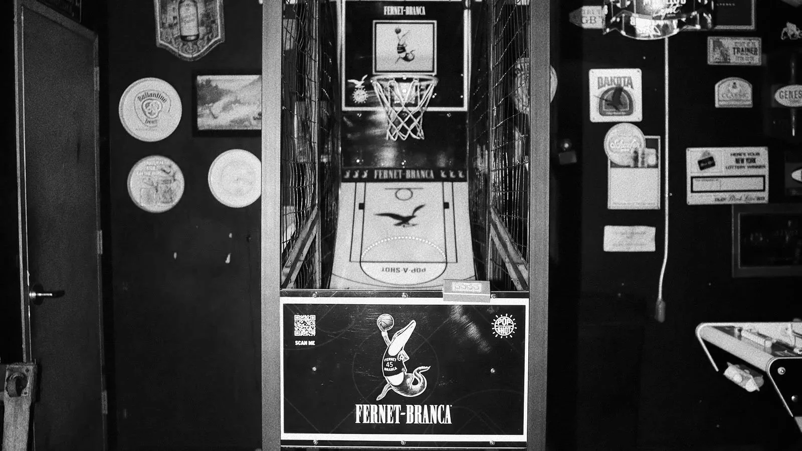 Black and white photo of a vintage arcade basketball game with a small basketball hoop, a painted court, and branding for Fernet-Branca. To the left, decorative wall plates and a framed picture, to the right, a coin slot and game controls. Part of a 