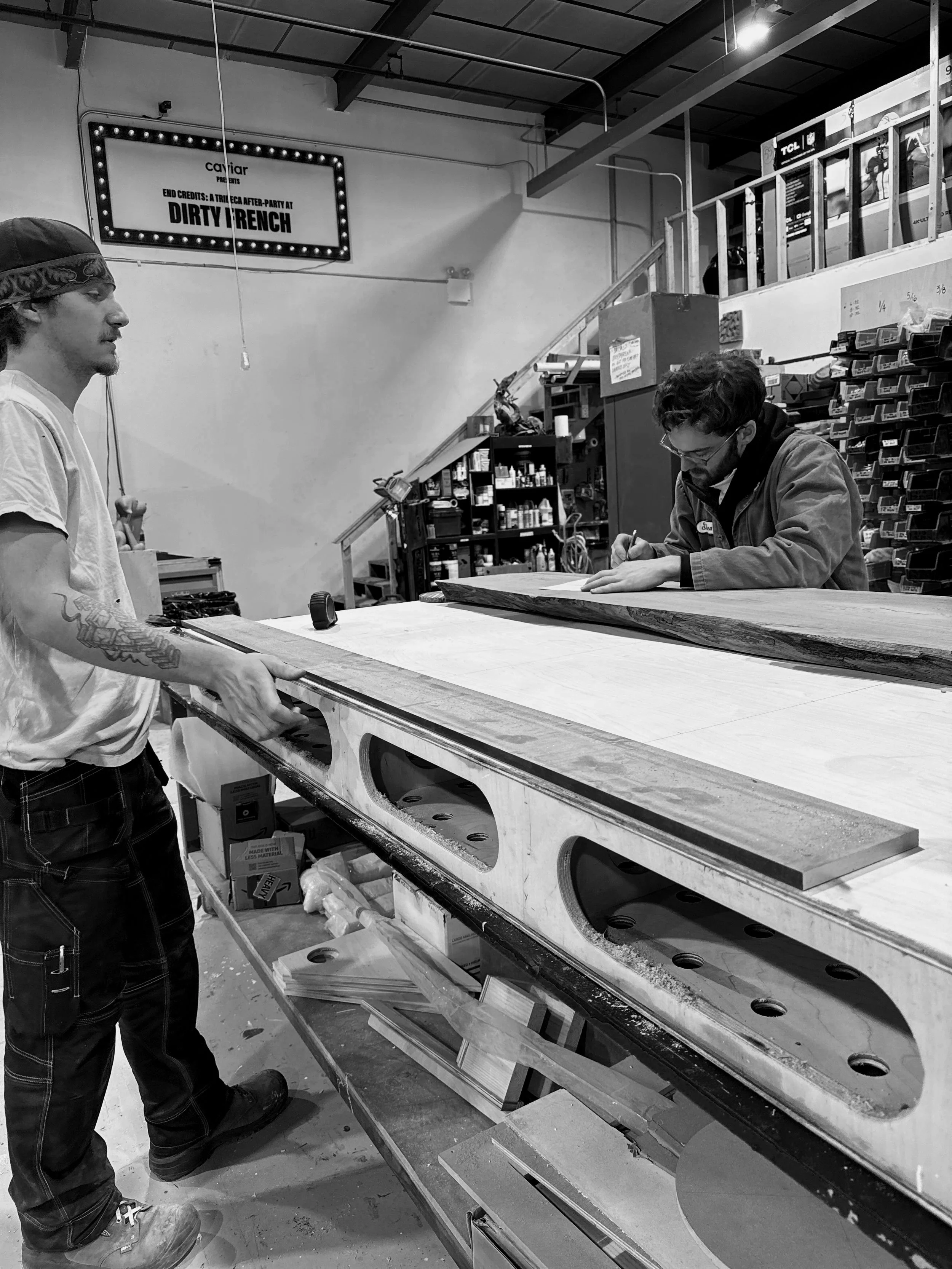 Two men working with a large piece of wood in a woodworking shop. One man is standing and holding the wood, the other man is sitting and writing. The workshop has tools, shelves. 