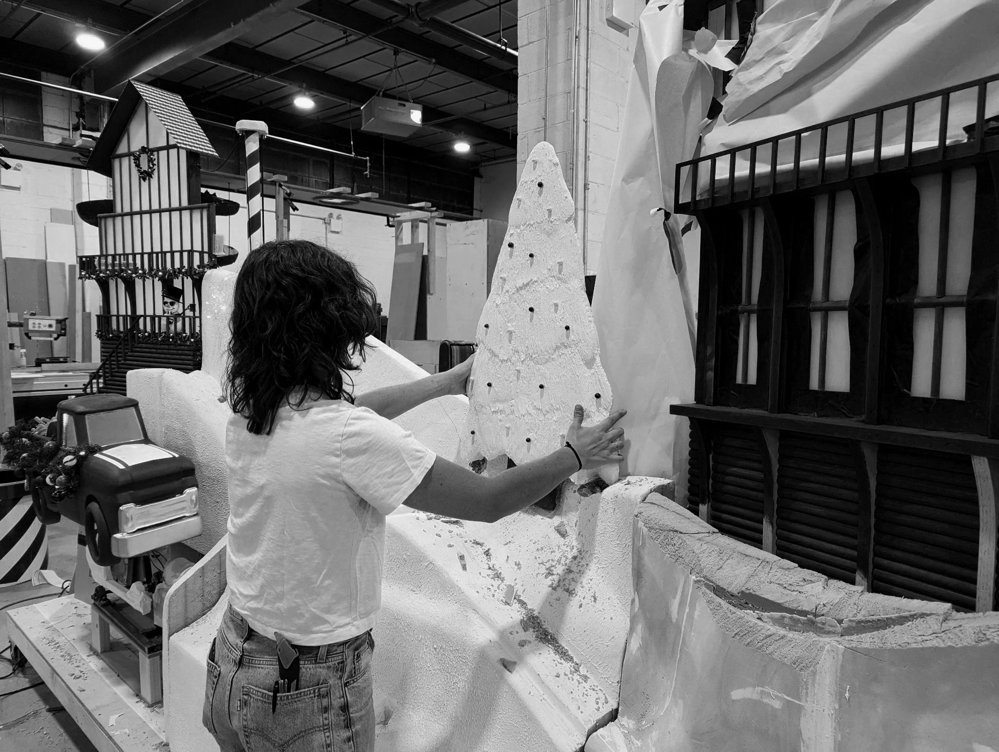 Woman assembling a large, decorated Christmas tree prop made of foam in a workshop, with other holiday decorations and structures in the background. Dry fitting a holiday window display 