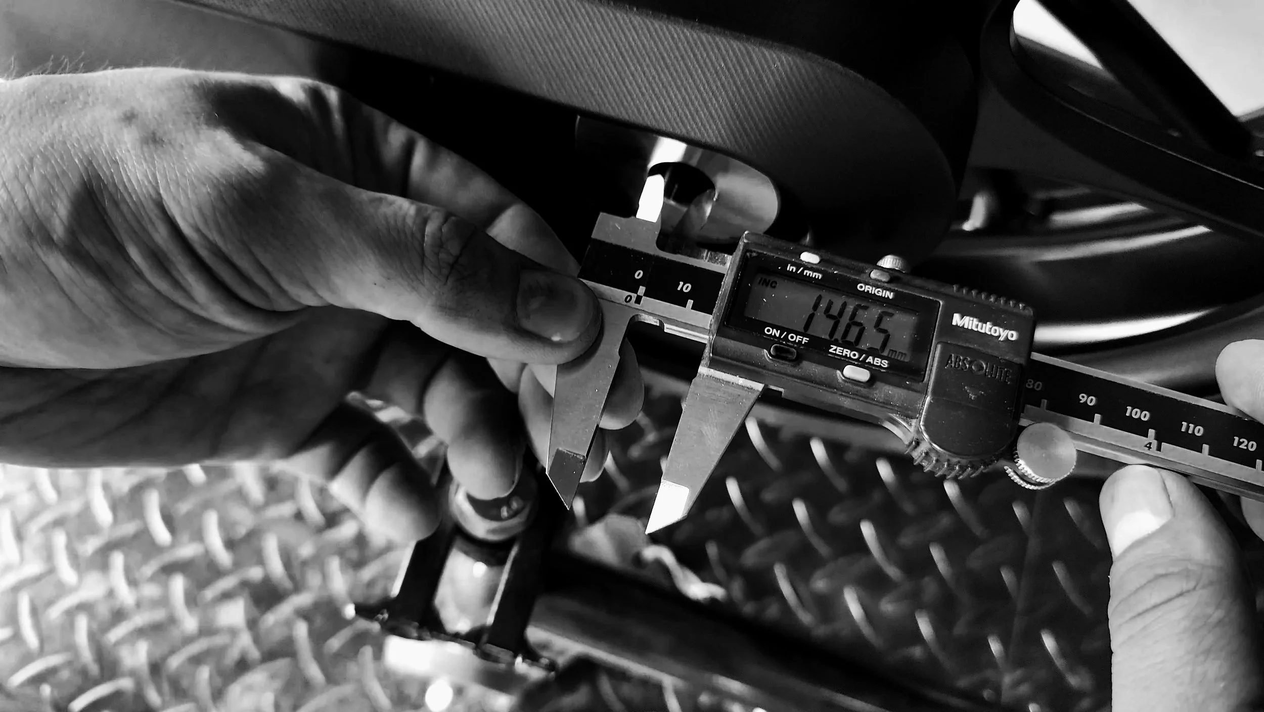 A person is using a digital caliper to measure the thickness or diameter of an object against a metal surface with a diamond plate pattern.