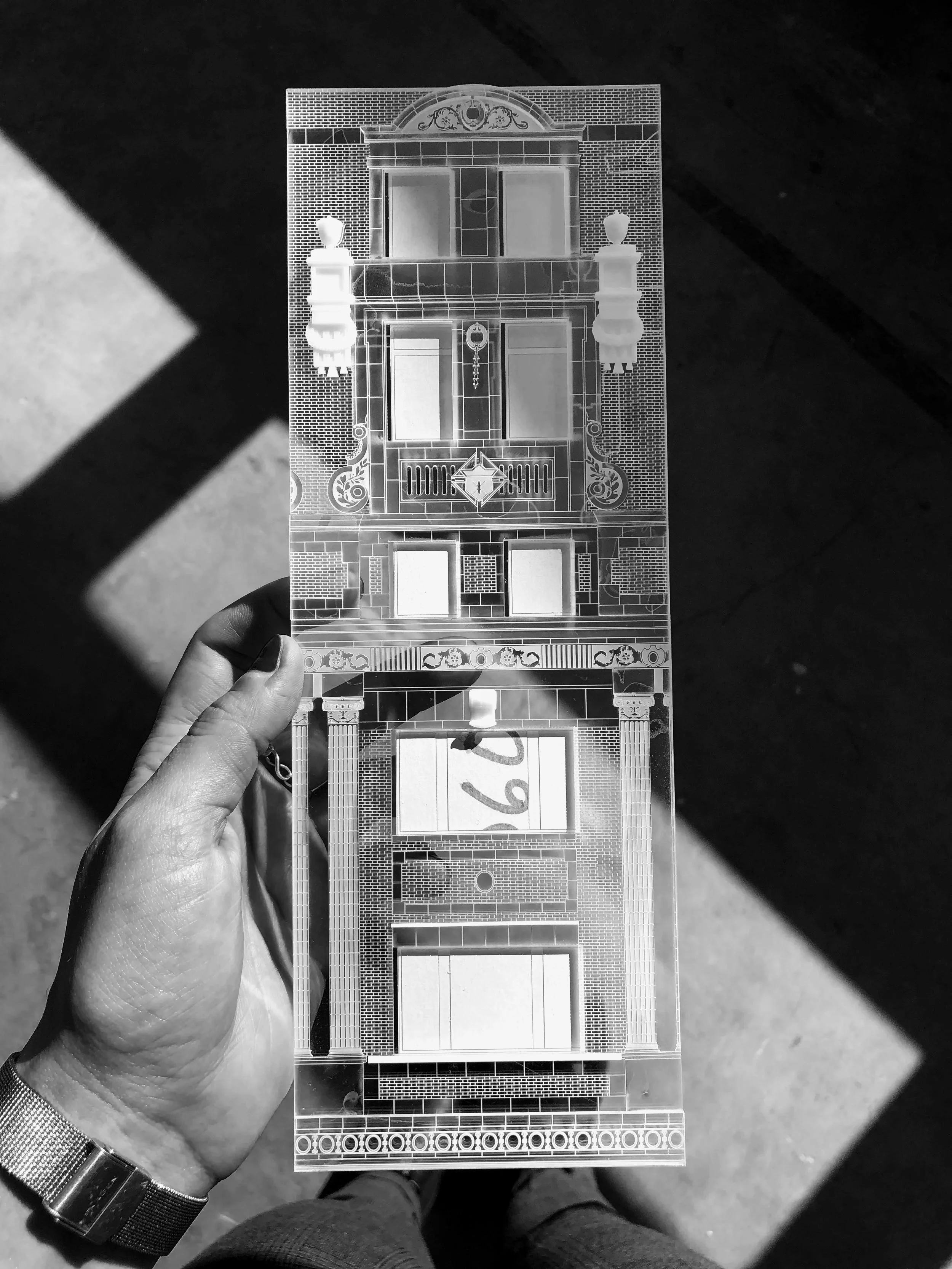 Hand holding a transparent acrylic sheet with architectural drawings, including a building facade with detailed windows, columns, and decorative elements. laser cut settings in clear acrylic for building details in miniature