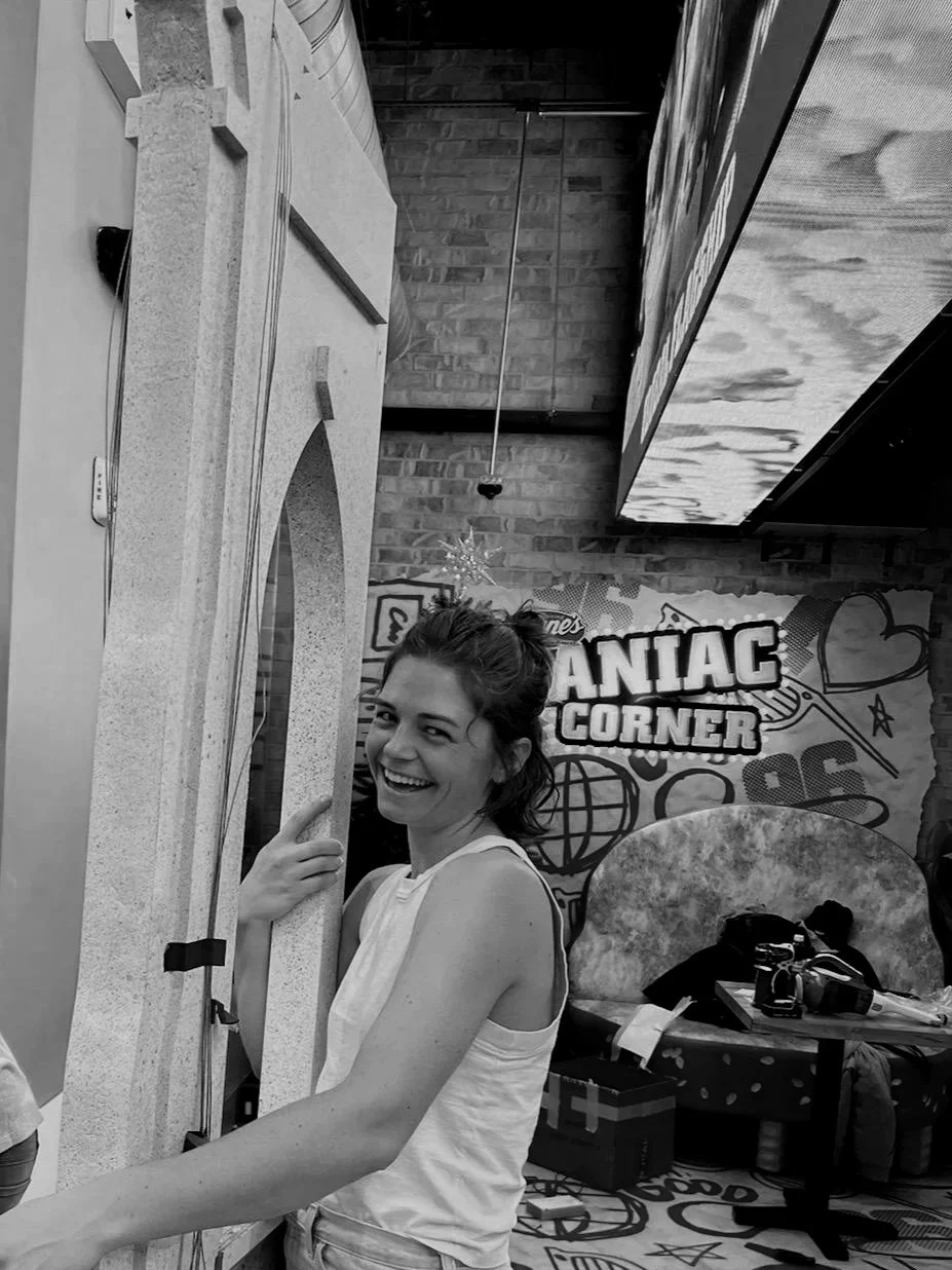 A smiling woman with her hair tied back, wearing a sleeveless top, standing in front of a photo booth at Raising Cane's Chicken Fingers, with a decorative wall featuring the words 'Caniac Corner' in the background.