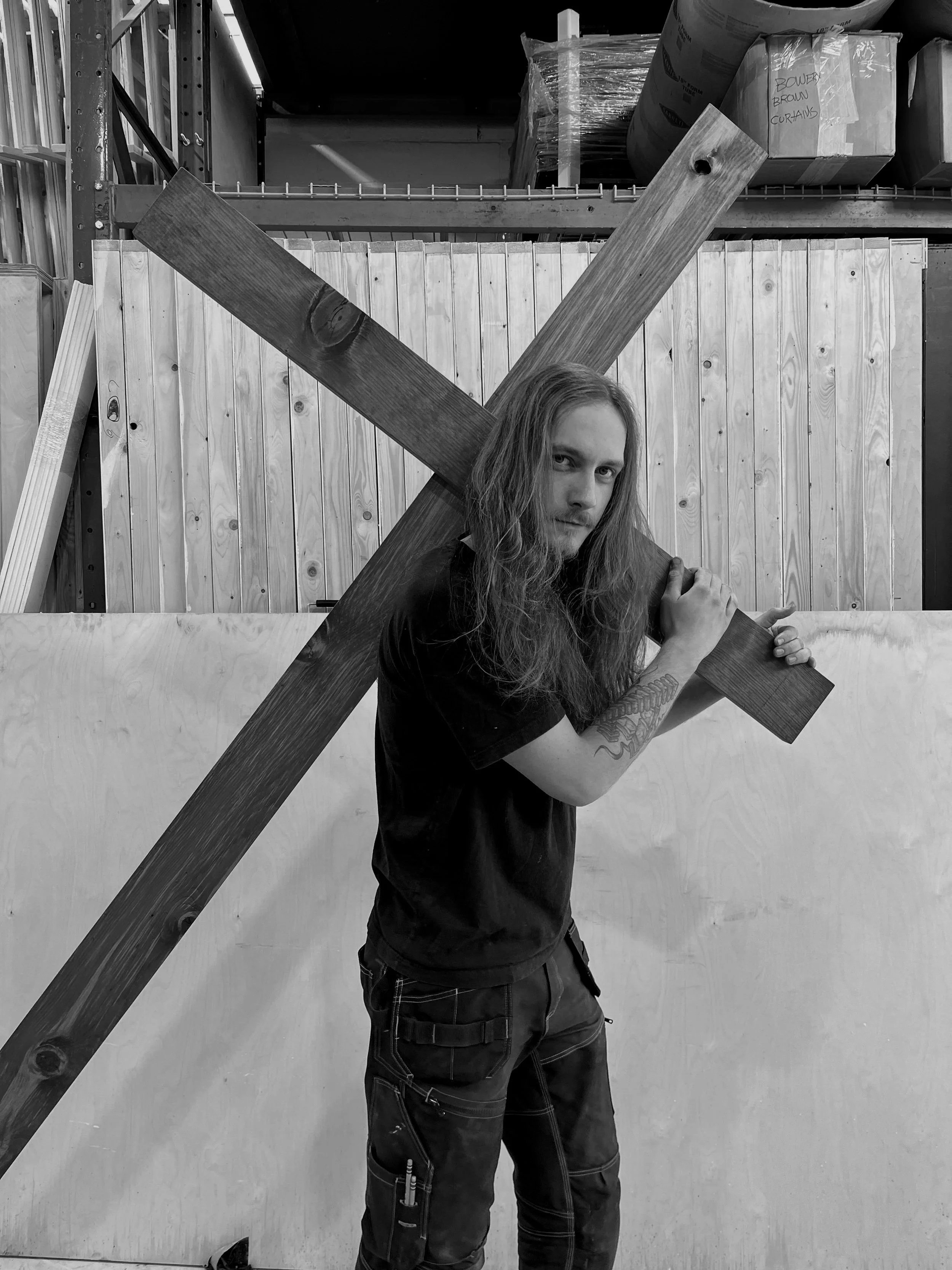 A man with long hair and tattoos holding a wooden cross on his shoulder, standing in front of a wooden panel in a warehouse or workshop.