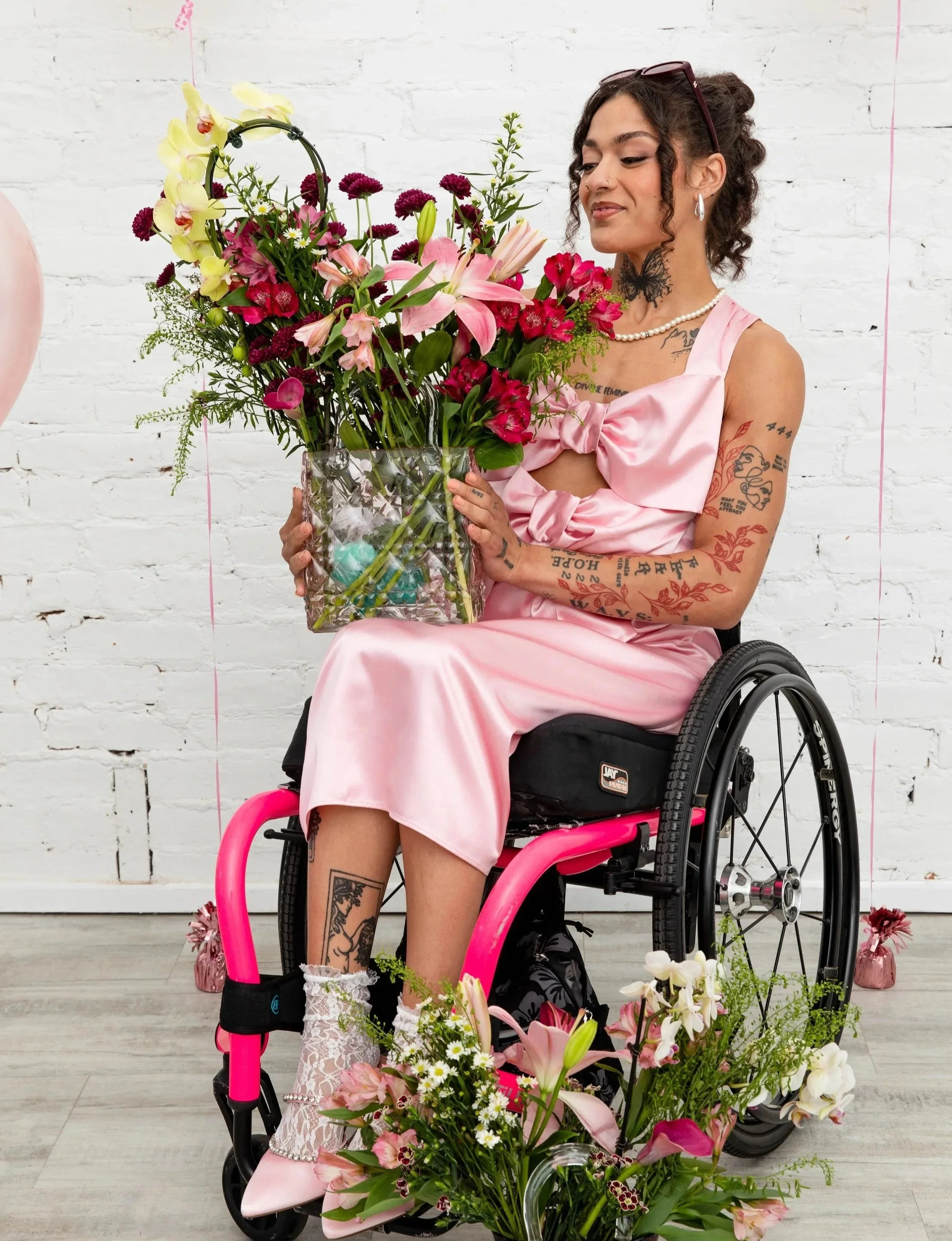 Woman in pink dress in wheelchair, holding a large bouquet of pink and burgandy flowers. Flowers are also on the ground around the wheelchair. White brick wall in the background.