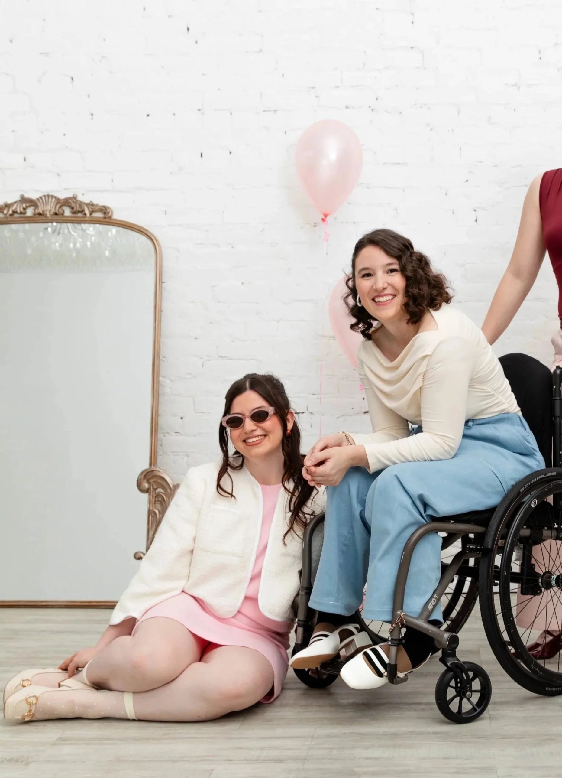 Group of women posing for a photo at a celebration, with pink balloons in the background, one woman in a wheelchair, smiling and wearing a cream top, pink skirt, and sunglasses, another woman kneeling on the floor with cream socks and shoes, and a th