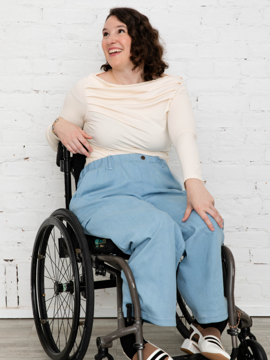 A smiling woman sitting in a wheelchair against a white brick wall, wearing a cream long-sleeve top and light blue jeans.