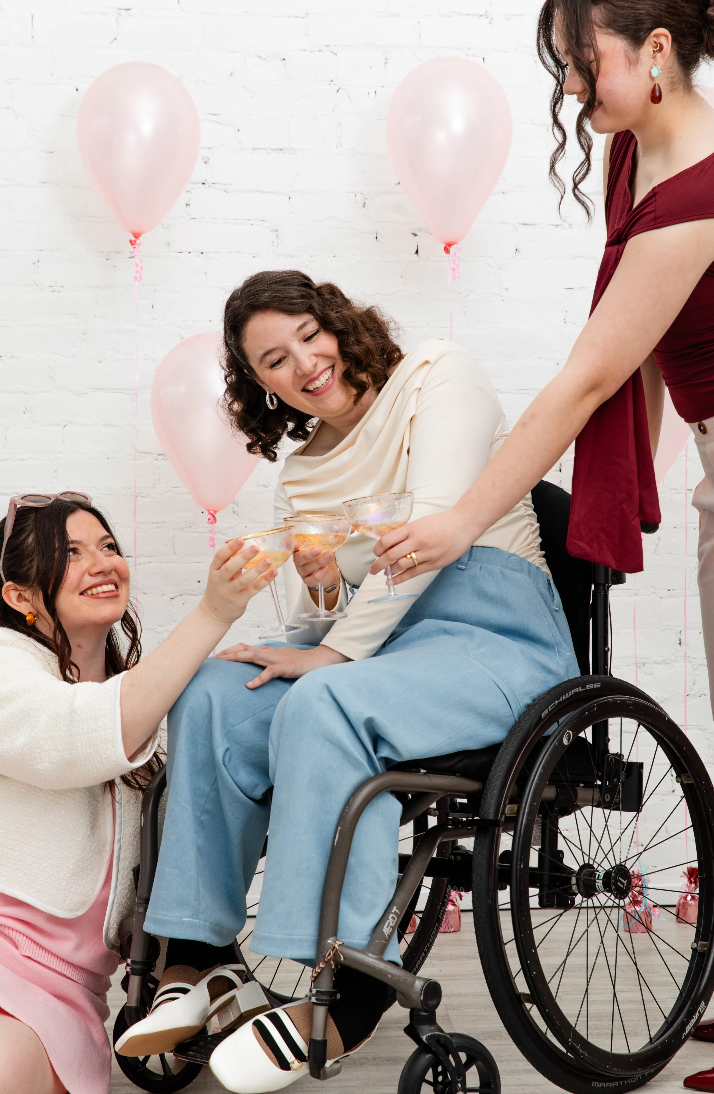 A woman in a wheelchair smiling and holding a glass while another woman with sunglasses holding a glass is kneeling beside her. A woman in a white outfit is standing next to the woman in the wheelchair. Pink balloons are in the background against a w