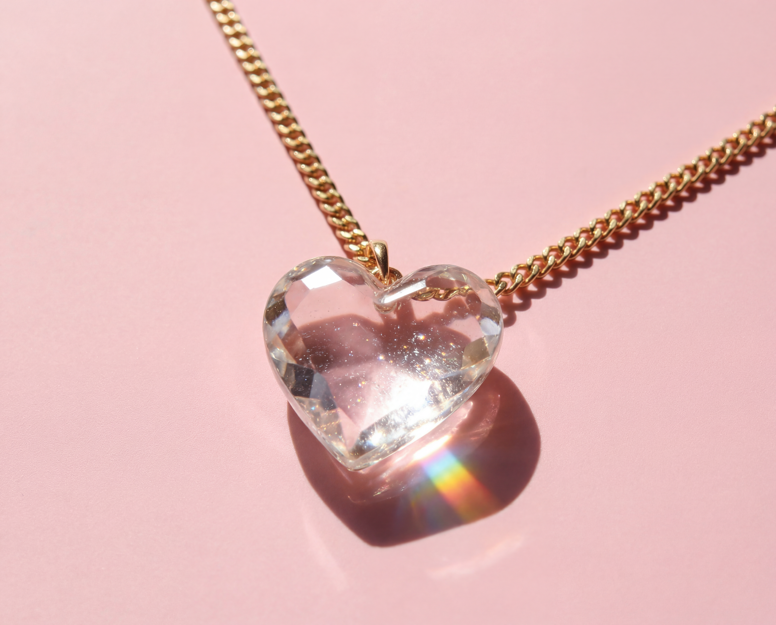 A gold chain necklace with a large, transparent heart-shaped pendant on a pink background.