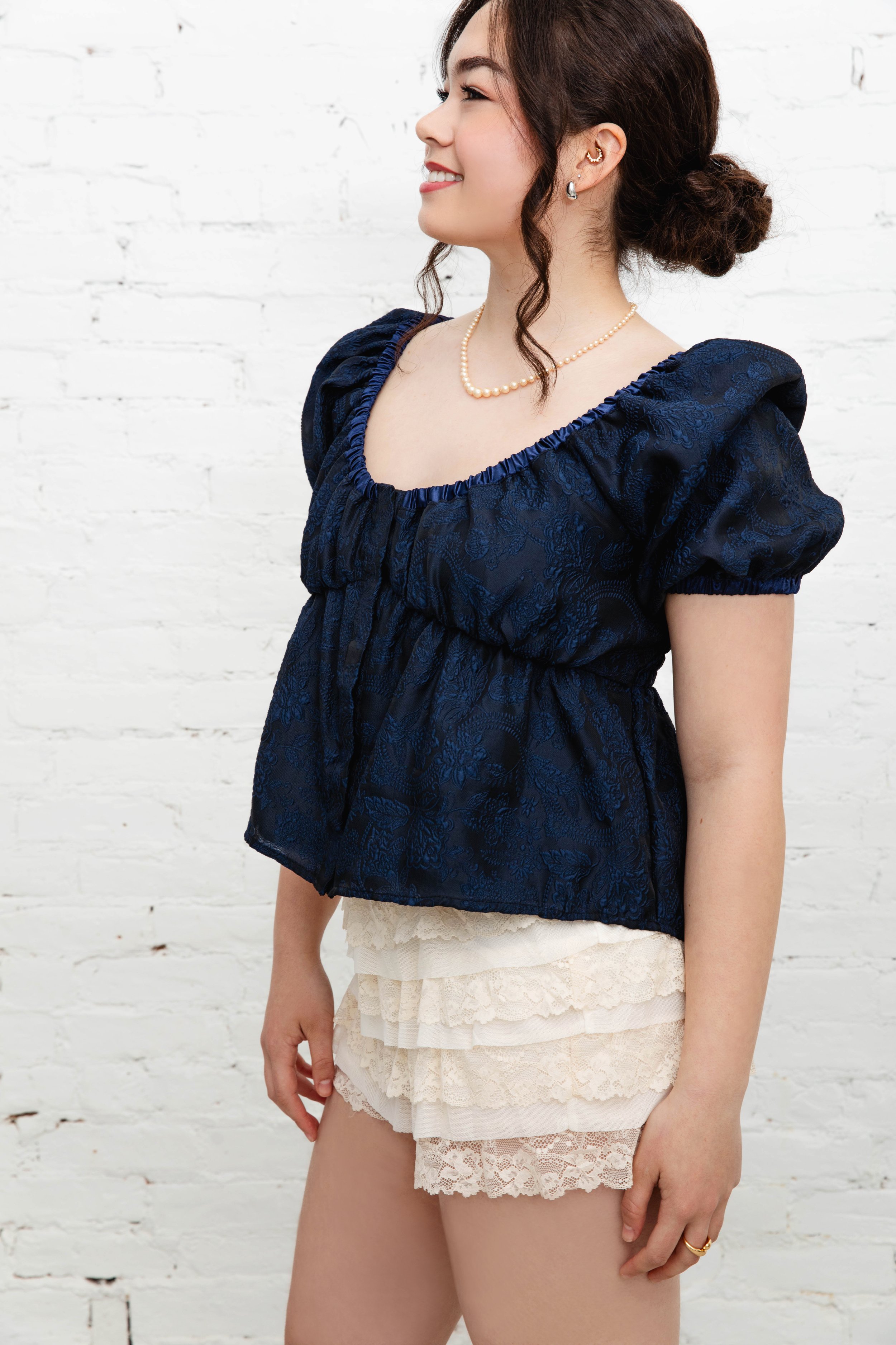 A woman in profile view smiling wearing a navy blue brocade top with puffy sleeves, lace ruffled shorts, a pearl necklace, earrings, and a gold ring, standing against a white brick wall. The blouse is shorter in the front and longer in the back.