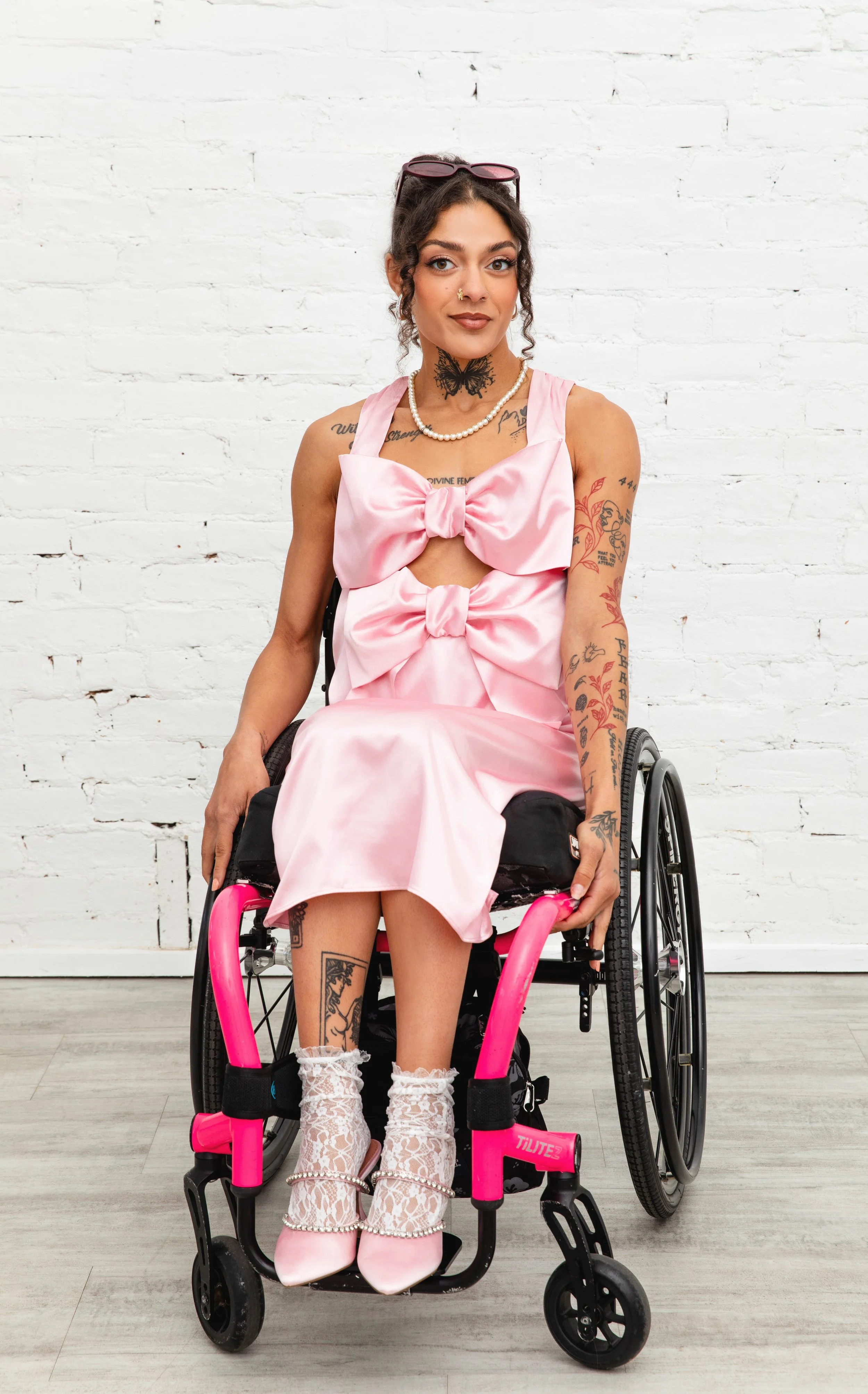 A woman with dark curly hair and tattoos wearing a pink dress with statement bows along the chest, white lace socks, and pink shoes sitting in a wheelchair against a white brick wall.