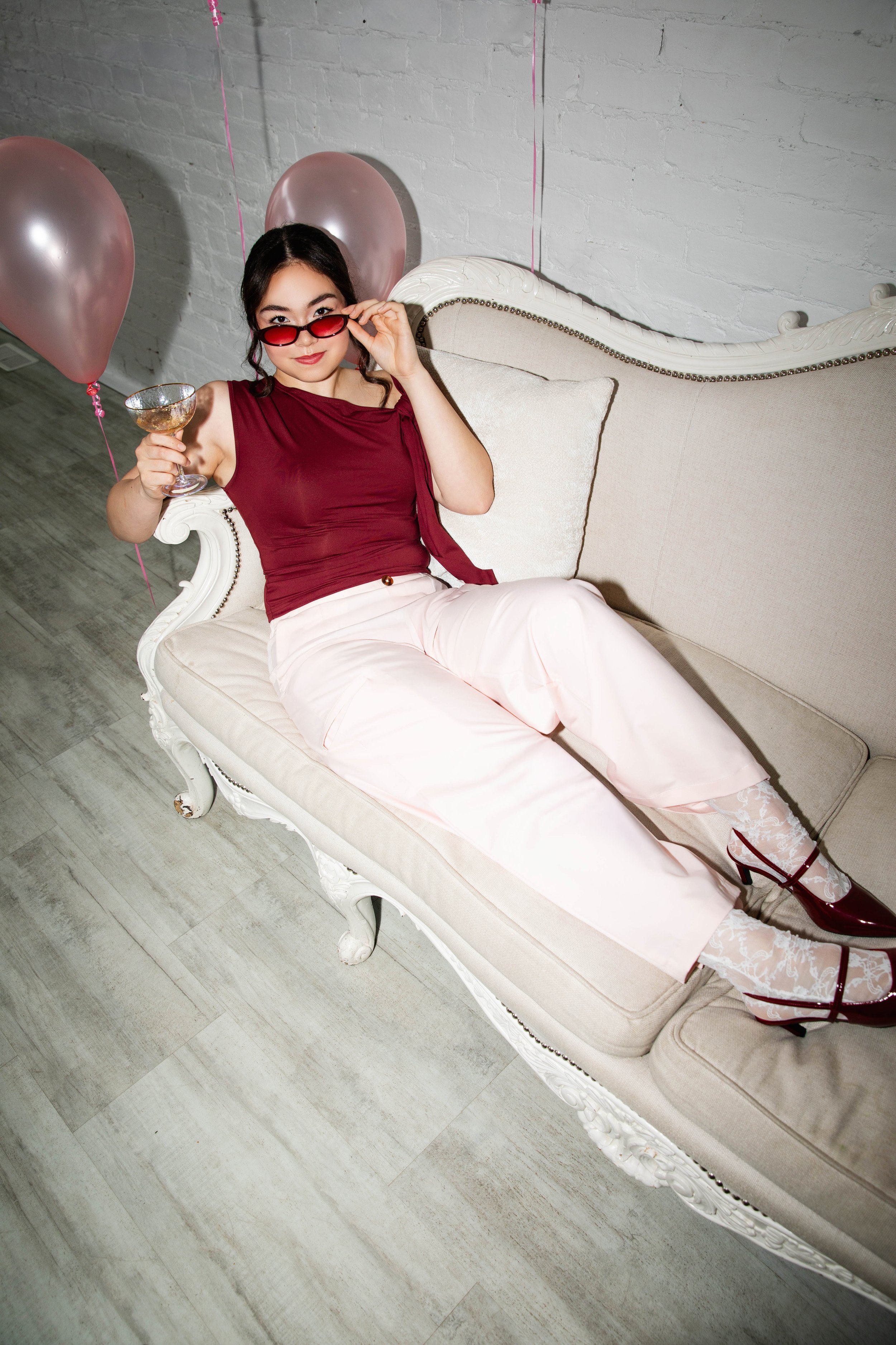 A woman sitting on a vintage cream-colored sofa with white carved wood detail, holding a glass of wine, wearing red sunglasses, a red sleeveless top, light pink pants, and high-heeled shoes with lace design, with pink balloons and pink streamers in t