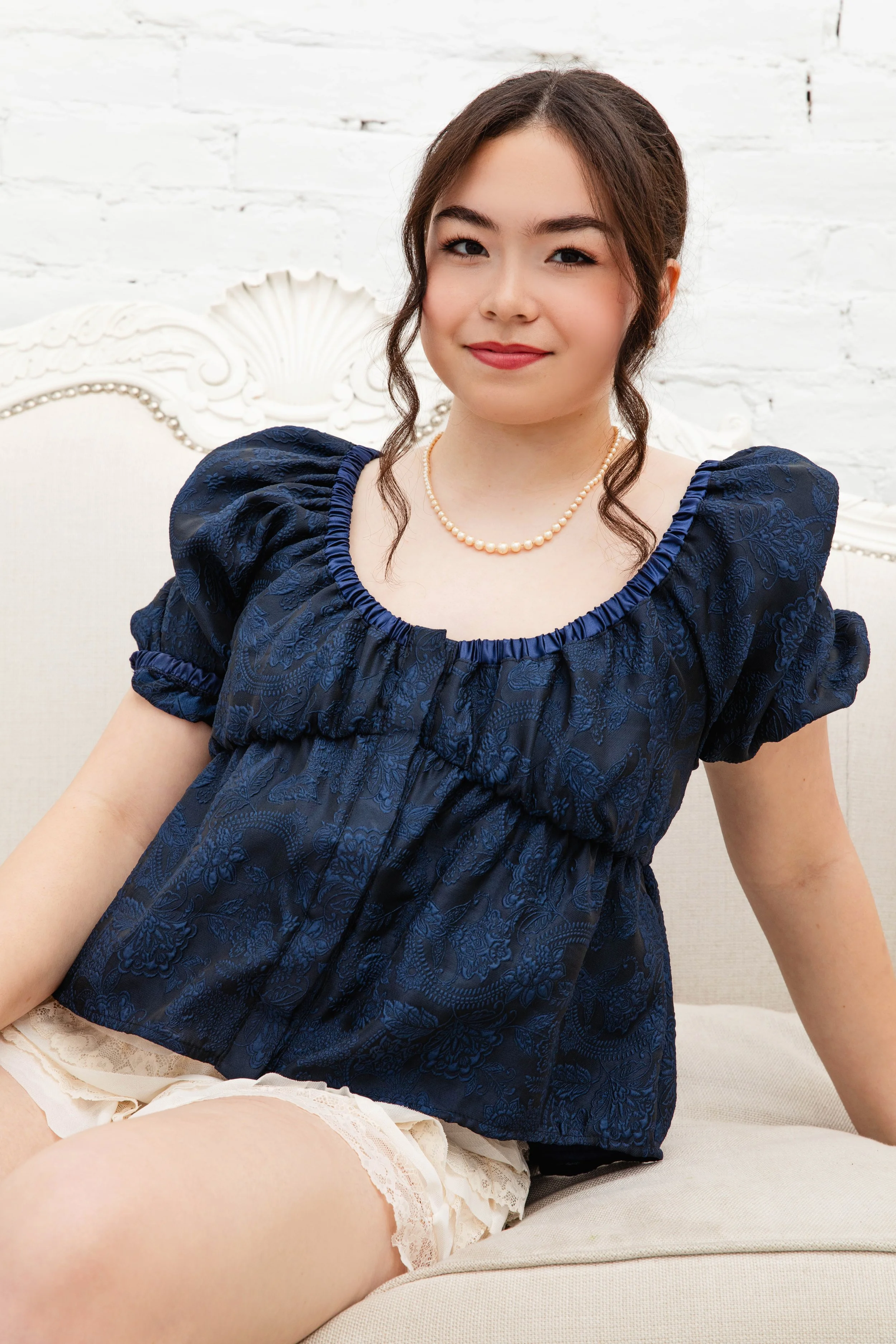 A woman with long dark hair in an updo, wearing a dark blue brocade blouse with puffed sleeves and pearl necklace, sitting on a white vintage-style sofa in front of a white brick wall, smiling gently at the camera.