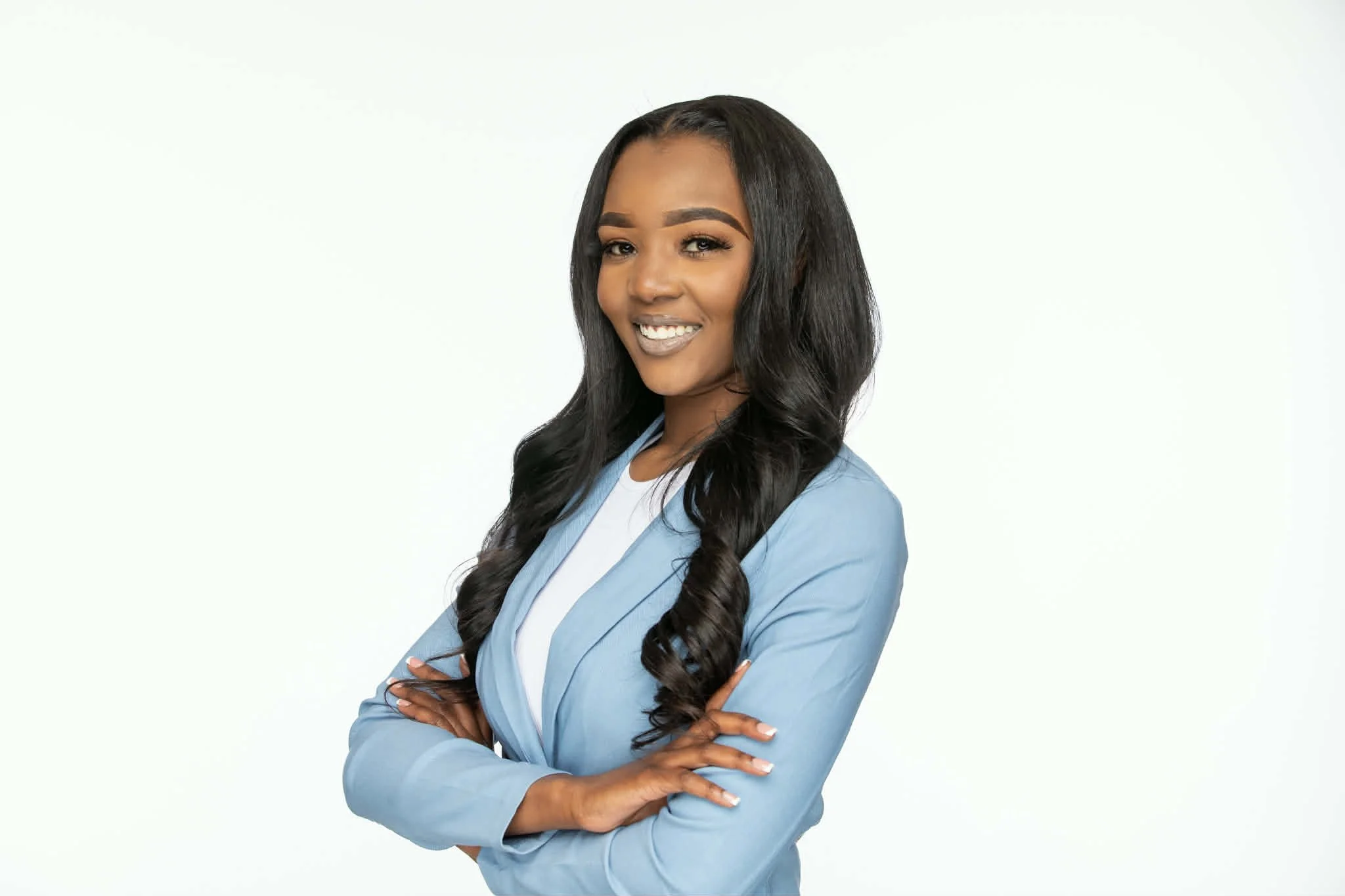 A confident African American woman with long curly black hair, wearing a light blue blazer over a white top, standing with arms crossed and smiling against a plain white background.